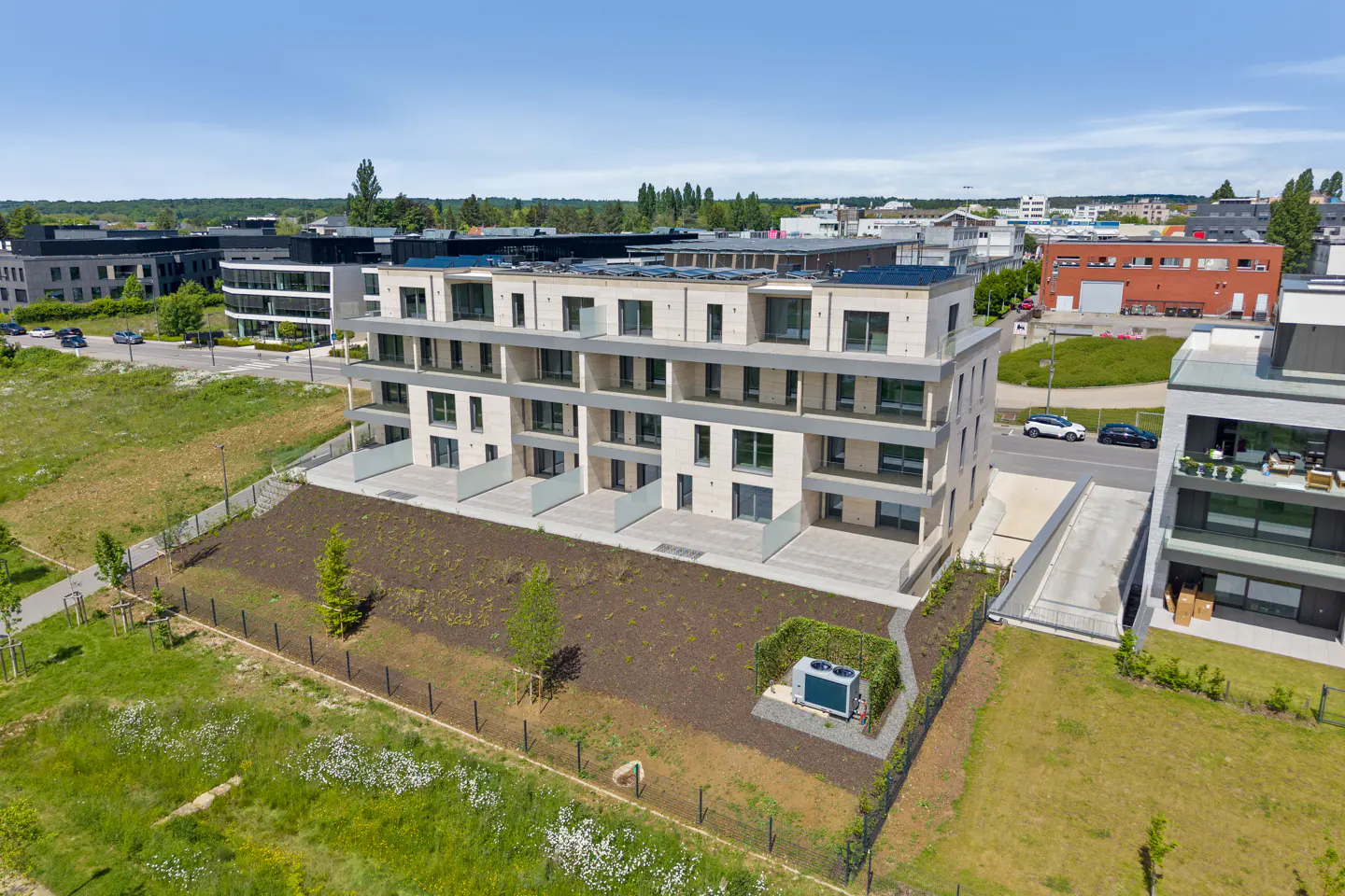 Aerial view of a modern, light-stone apartment building with balconies, surrounded by green lawns and other buildings under a blue sky.