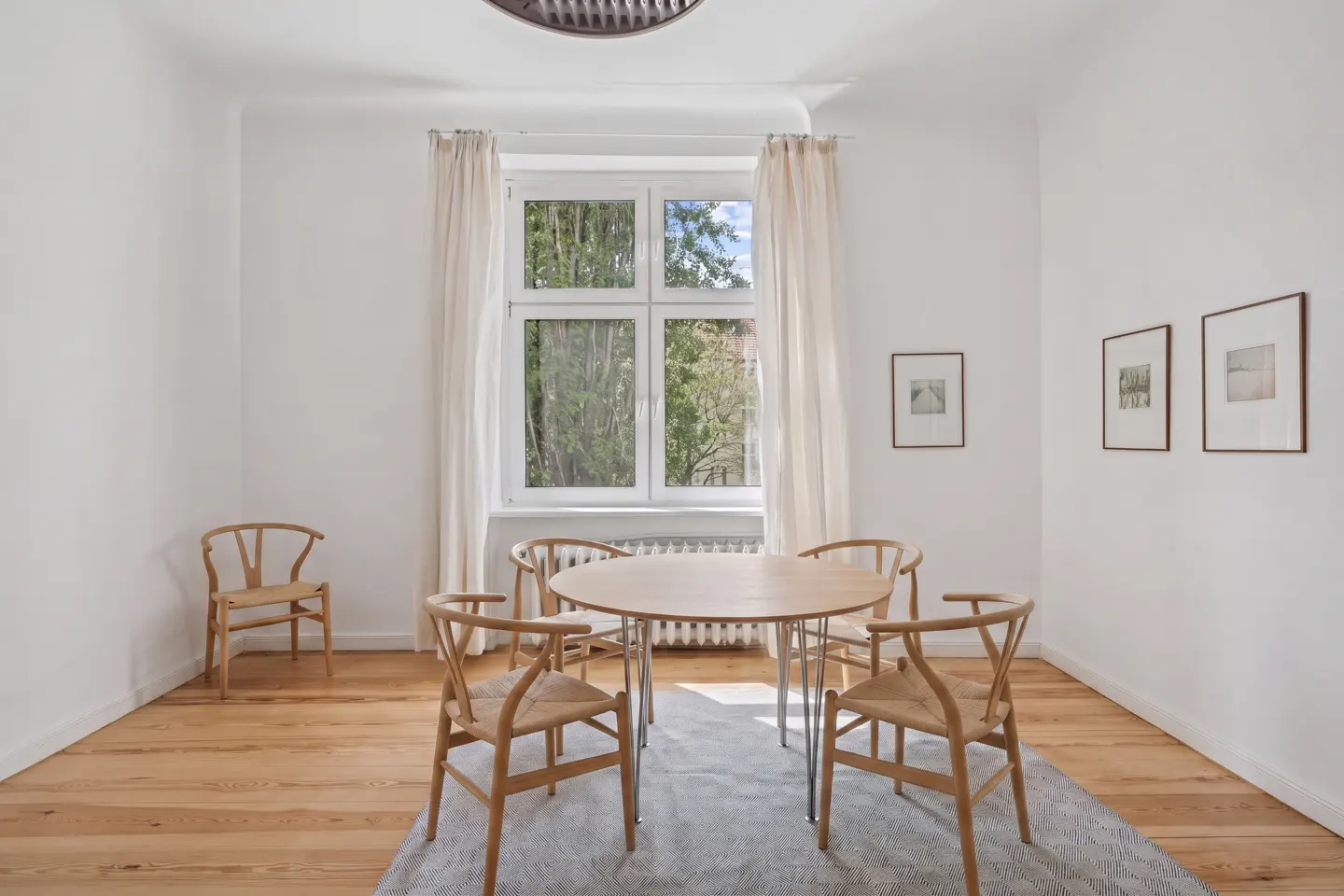 Bright dining room with white walls, wood floors, and a round table with six wishbone chairs on a gray rug. Three framed pictures hang on the wall.