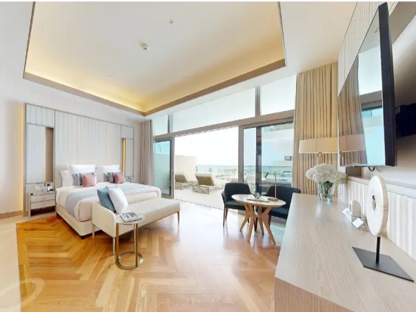 Bright, modern bedroom with a king bed, balcony with lounge chairs, and a small table with two chairs. The floor is wood and the walls are white.