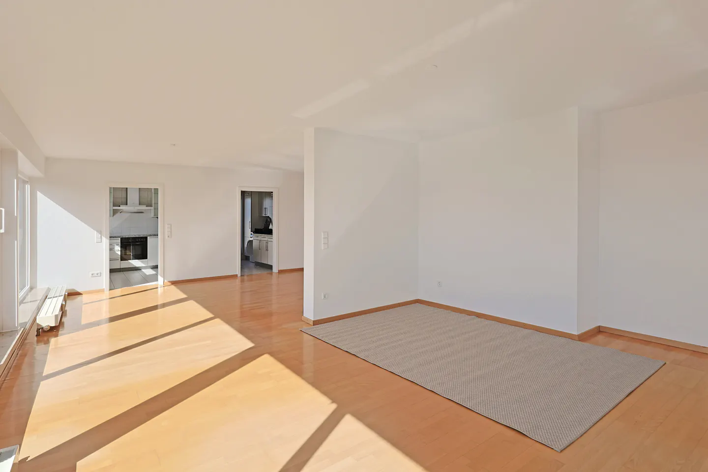Bright, empty room with hardwood floors, white walls, and a gray rug. Sunlight streams in from the left, illuminating the floor. Kitchen visible through doorway.