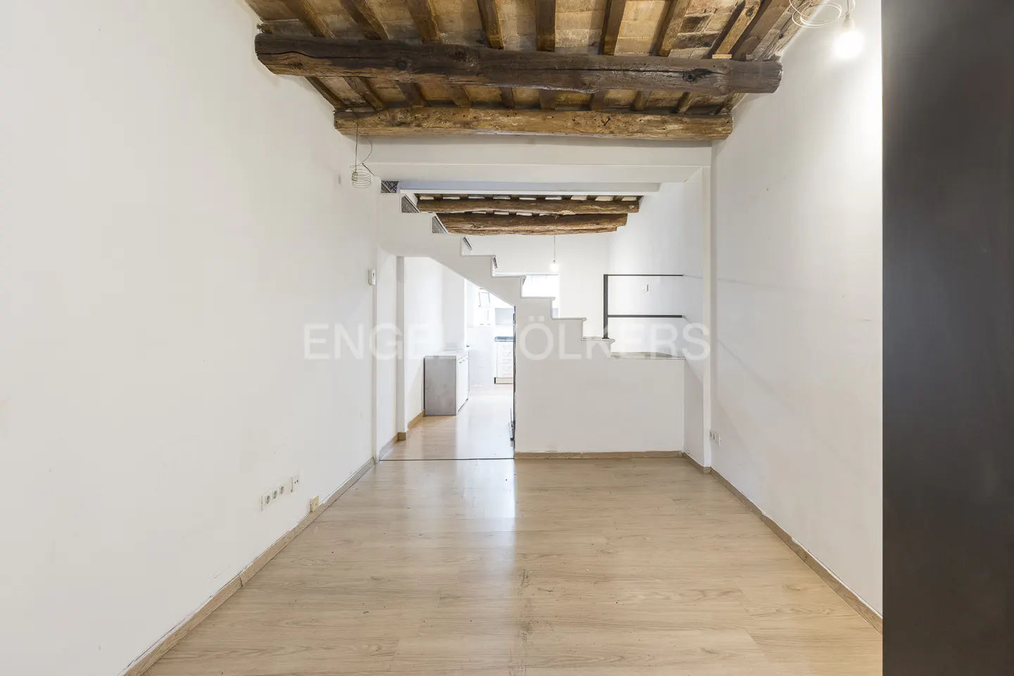 Bright, empty room with wood floors, white walls, and exposed wood beams on the ceiling. Stairs lead to another room.
