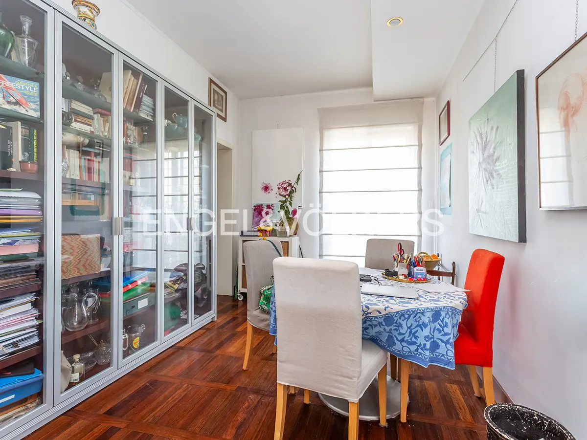 Bright dining room with a large glass-door bookcase, a round table with blue floral tablecloth, and chairs in gray and red. Artwork hangs on the white walls.