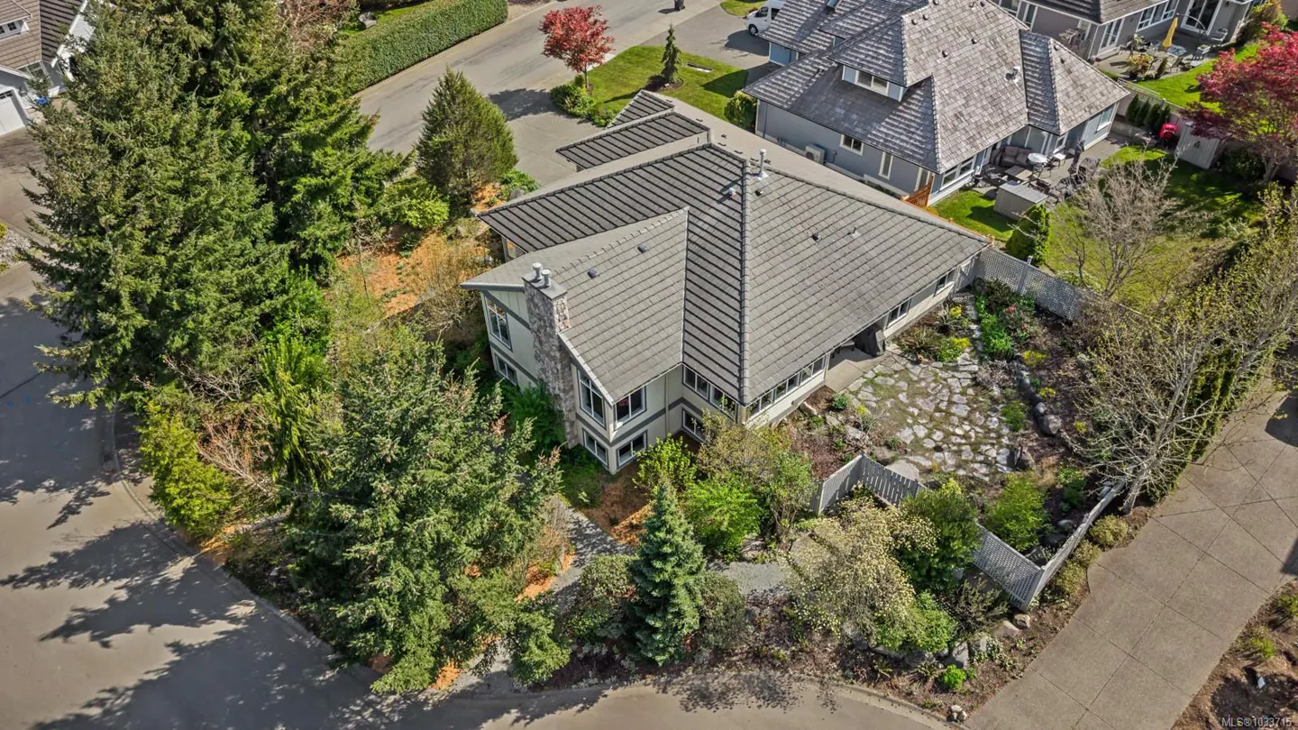 Aerial view of a two-story house with a gray tiled roof, stone chimney, and lush green landscaping. A stone pathway leads to the house.