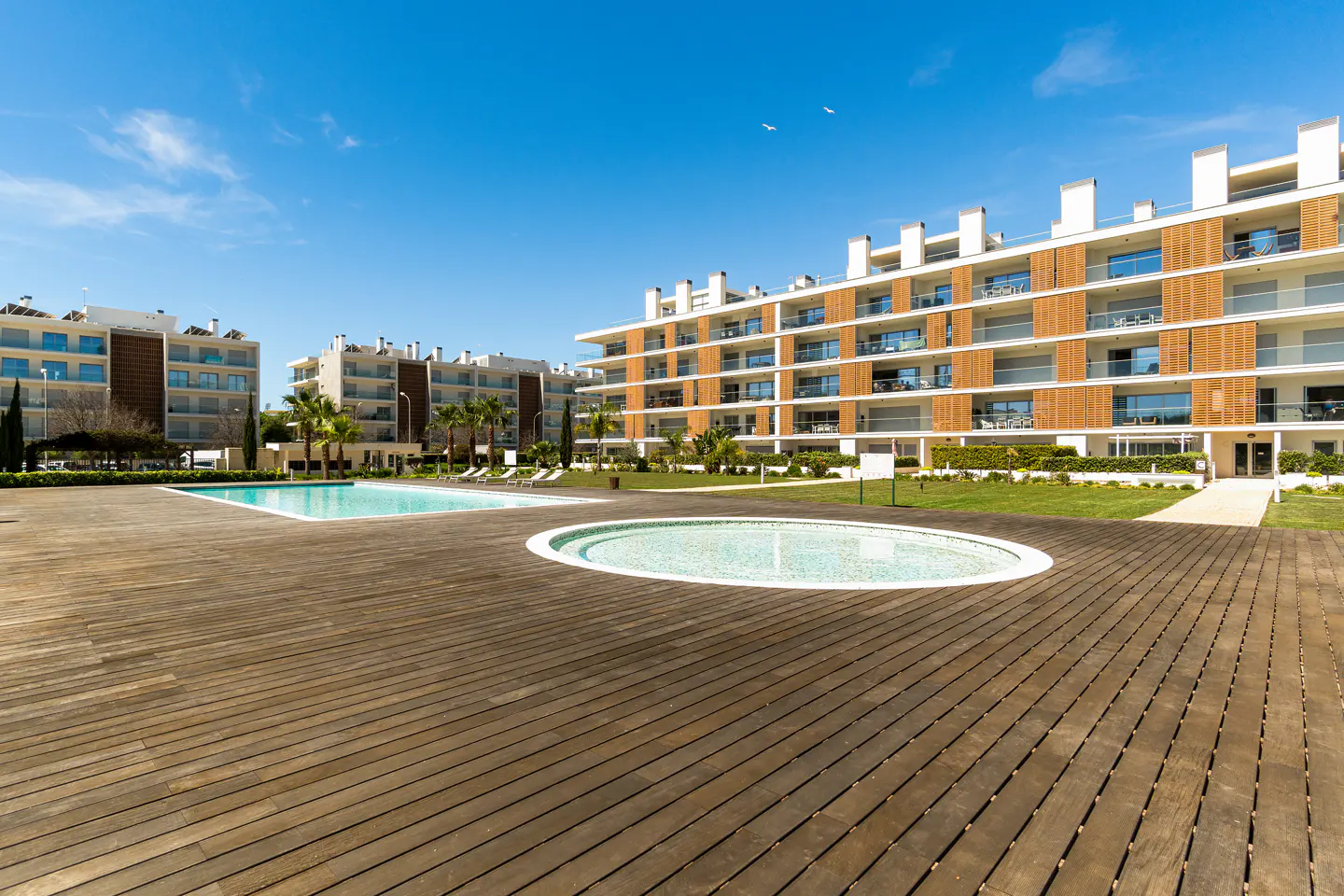 Exterior view of modern apartments with a pool and wooden deck under a blue sky.