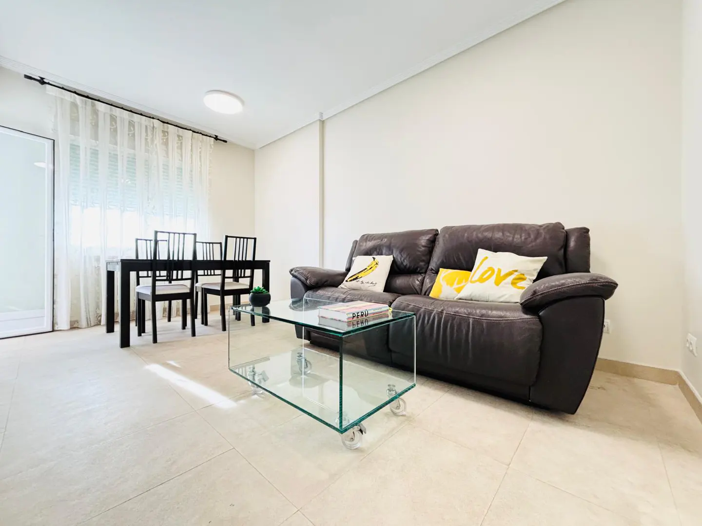 Living room with a brown leather sofa, glass coffee table, and black dining table with chairs. Cream-colored walls and tile flooring.