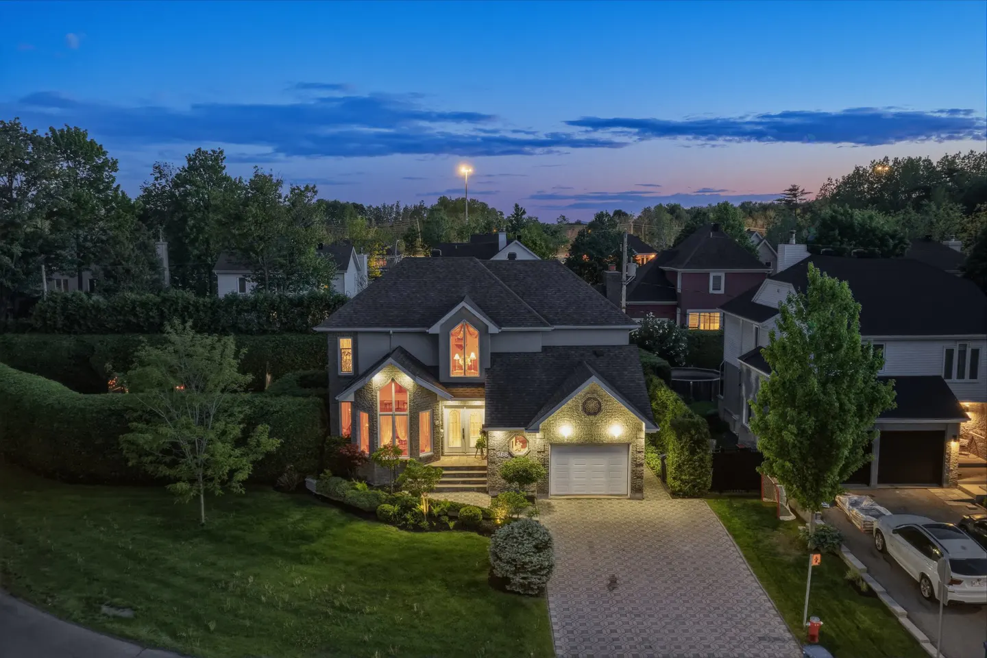 Two-story suburban home with stone facade, lit windows, and paved driveway at dusk, surrounded by trees and green lawn.