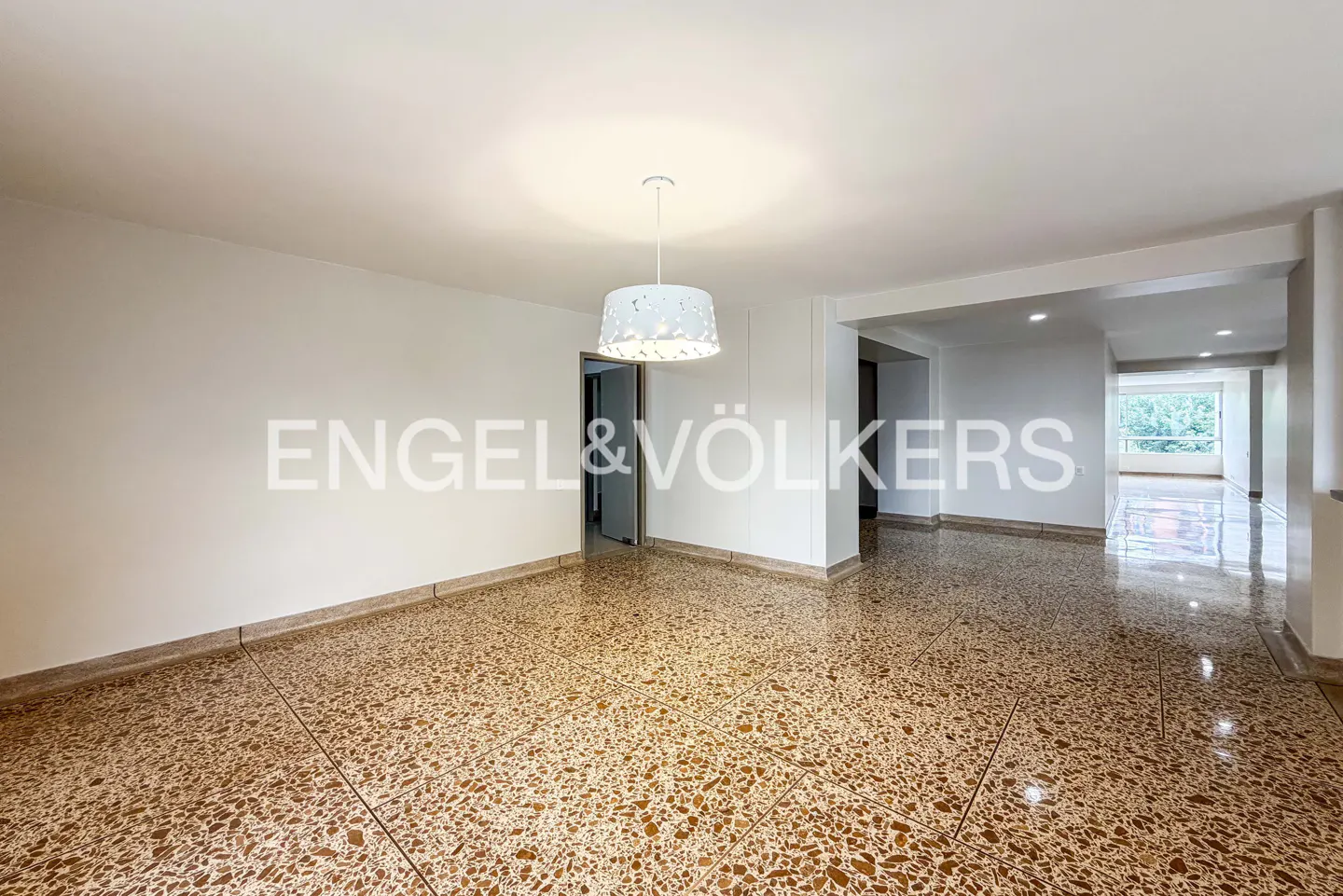 Empty room with white walls, speckled brown floor, and a white drum pendant light. Engel & Völkers logo is visible.