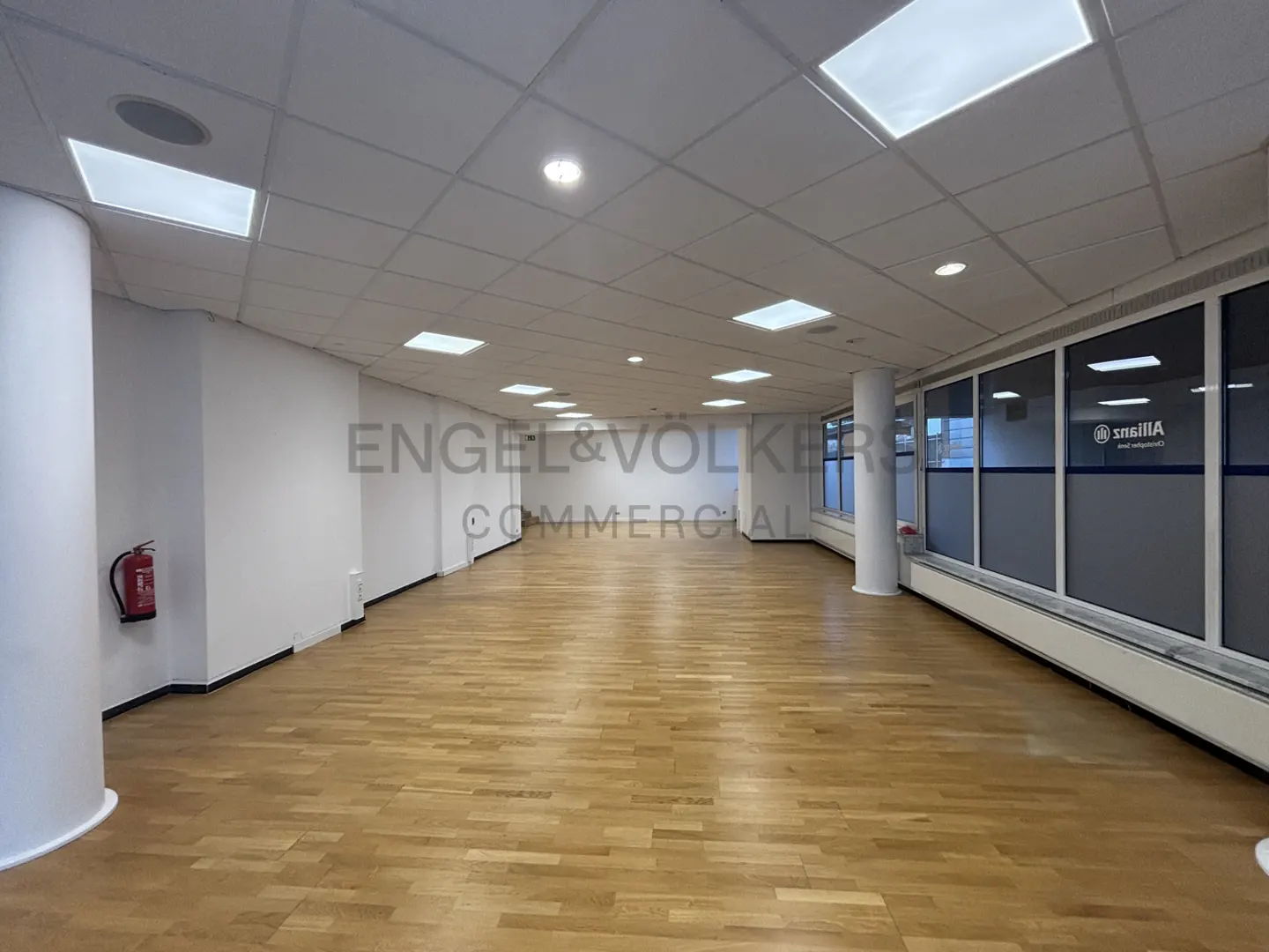 Bright, empty commercial space with wood floors, white walls, and a tiled ceiling with recessed lighting. Windows line the right wall. A fire extinguisher is visible on the left.