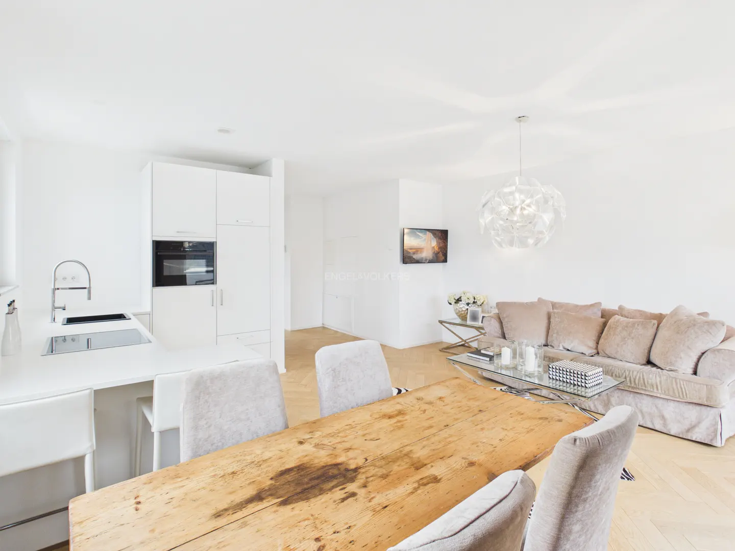 Bright, open-concept living space with a rustic wood table, white kitchen, and beige sofa. A modern chandelier hangs above the seating area.