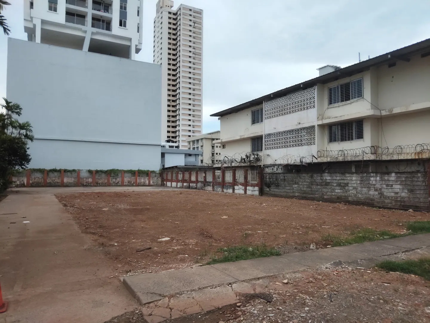 Vacant lot with red dirt, surrounded by a low wall and buildings. A tall white building is in the background.