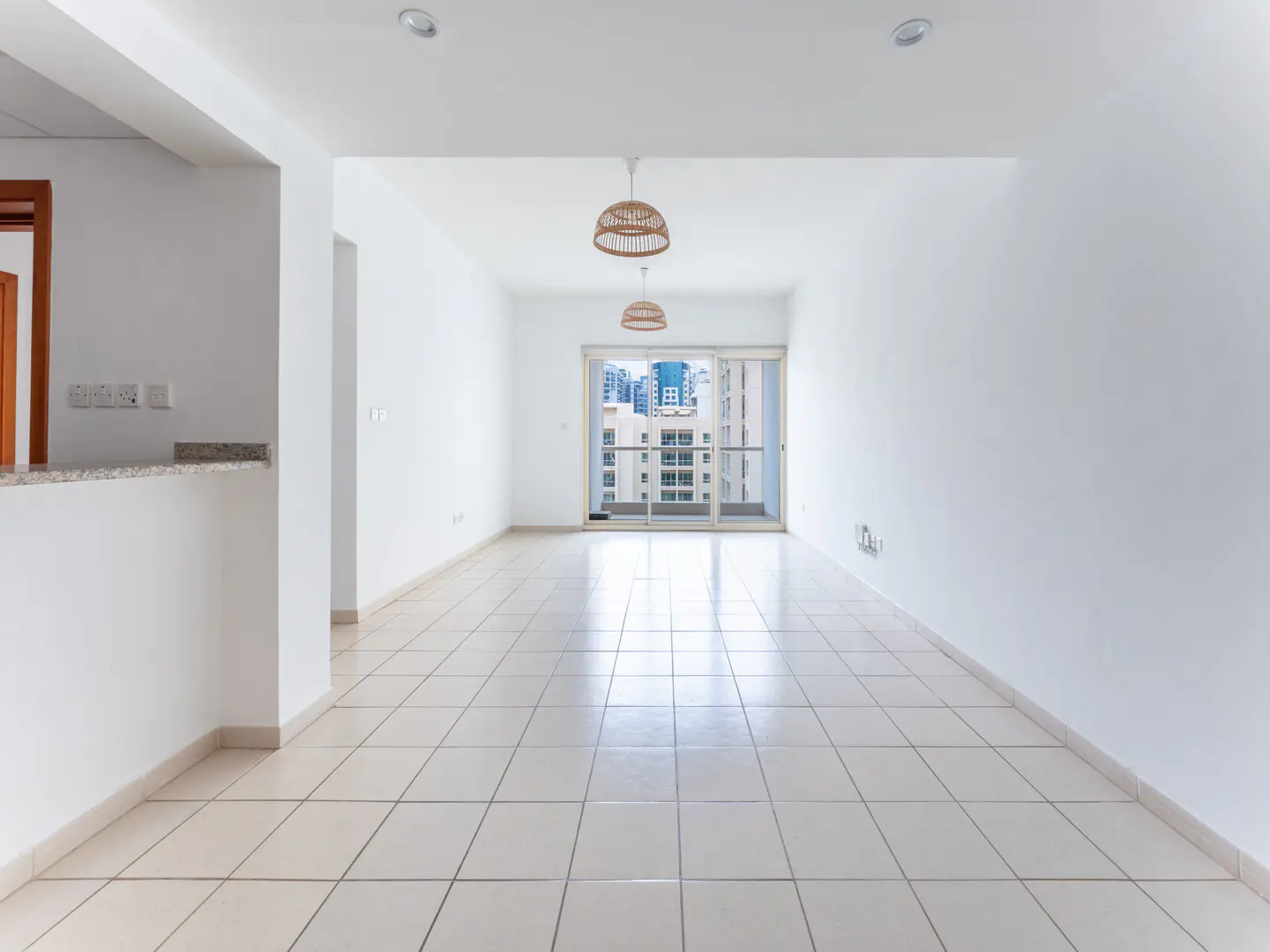Bright, empty room with white walls and tiled floor. Two wicker pendant lights hang from the ceiling. Balcony doors offer a city view.