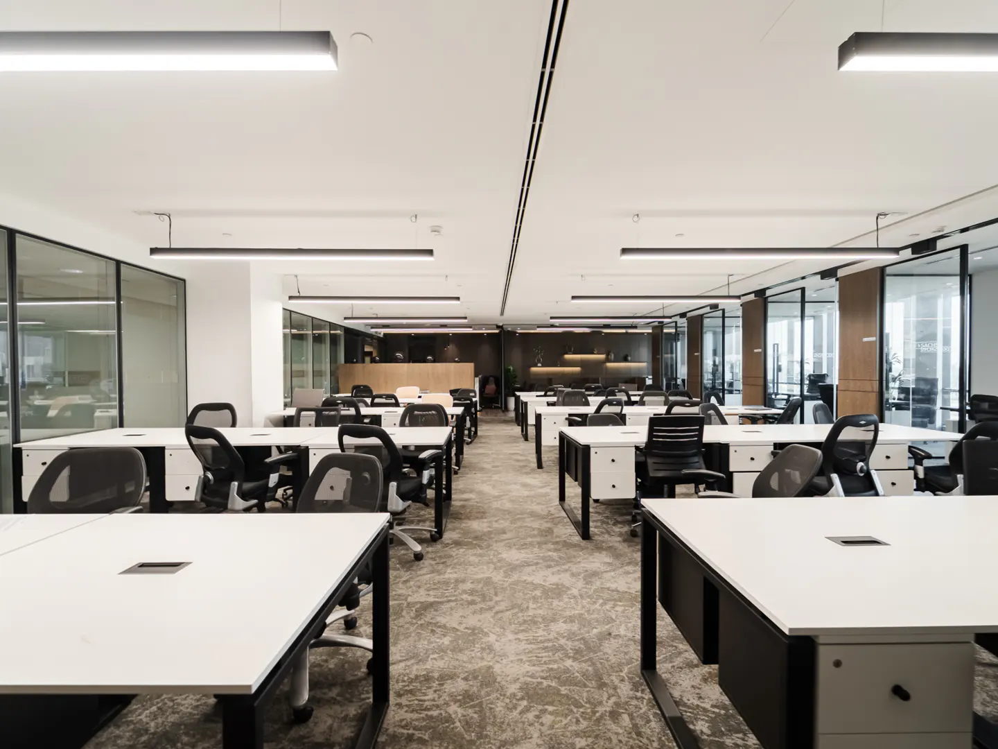 Bright, modern office space with rows of white desks, black chairs, and gray carpet. Glass-walled offices line the perimeter.