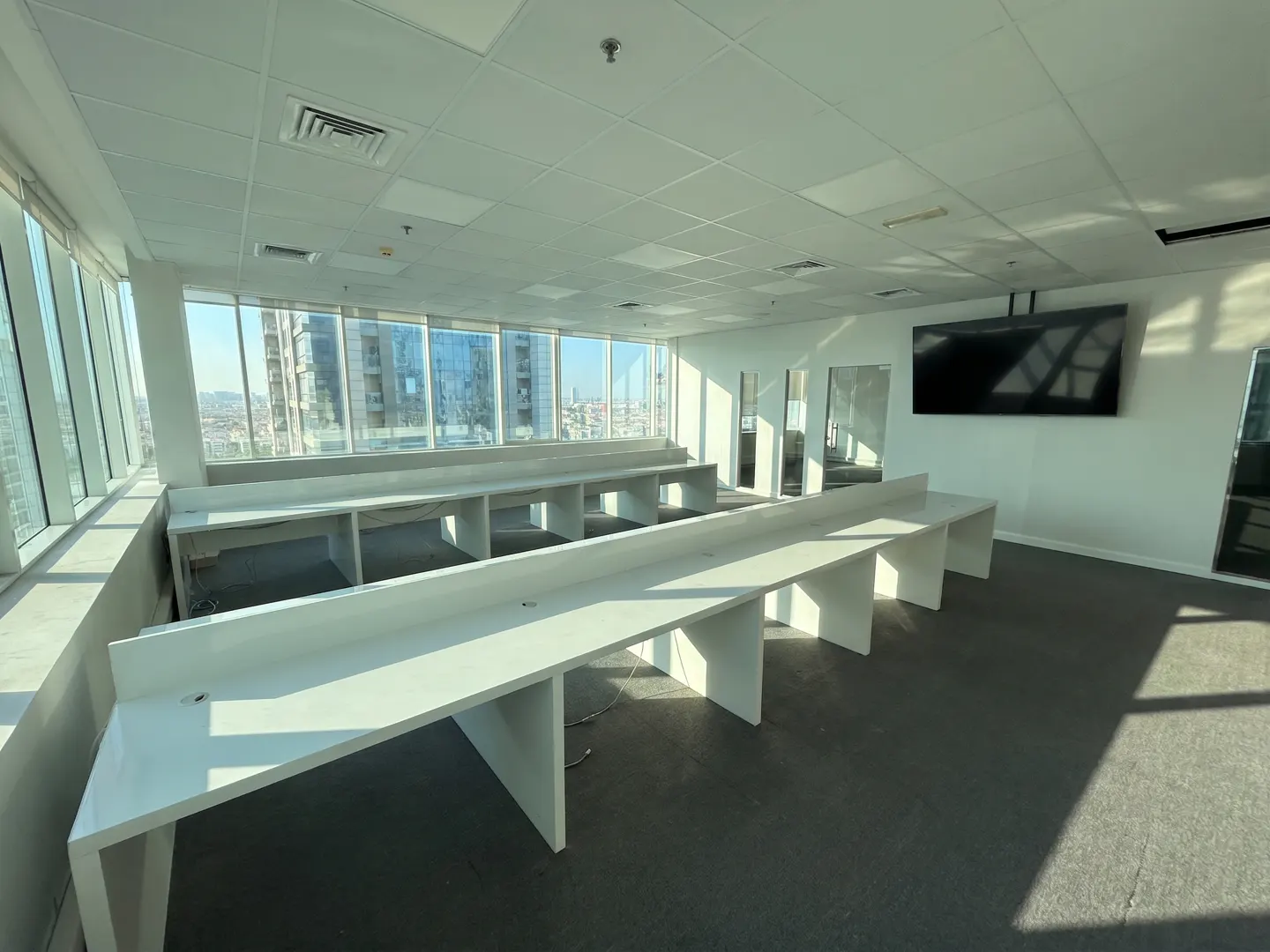 Bright, empty office space with long white desks, gray carpet, and large windows overlooking a city. A black TV is mounted on the wall.