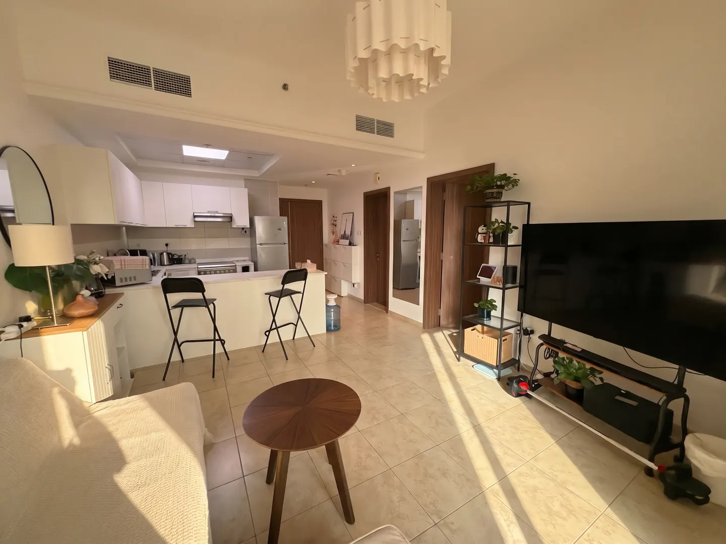 A bright, open-concept apartment with a kitchen, living area, and tile flooring. White cabinets, black stools, a round wood table, and a large TV are visible.