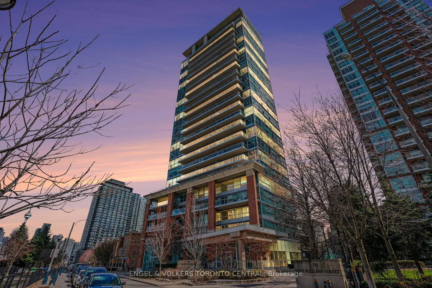 Exterior view of a tall, modern condo building with glass balconies at dusk. Bare trees frame the building against a pink and purple sky.