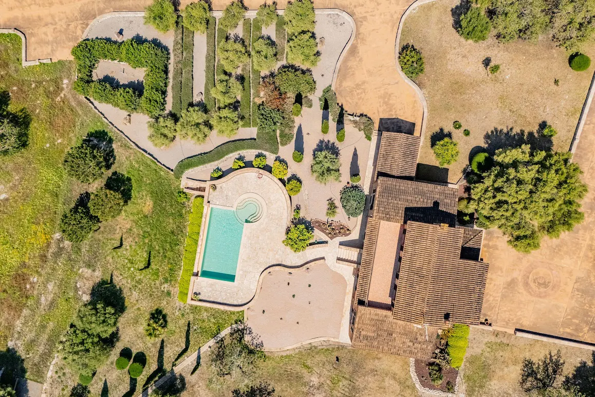 Aerial view of a luxury home with a brown tile roof, a turquoise pool, and manicured green gardens on a sunny day.