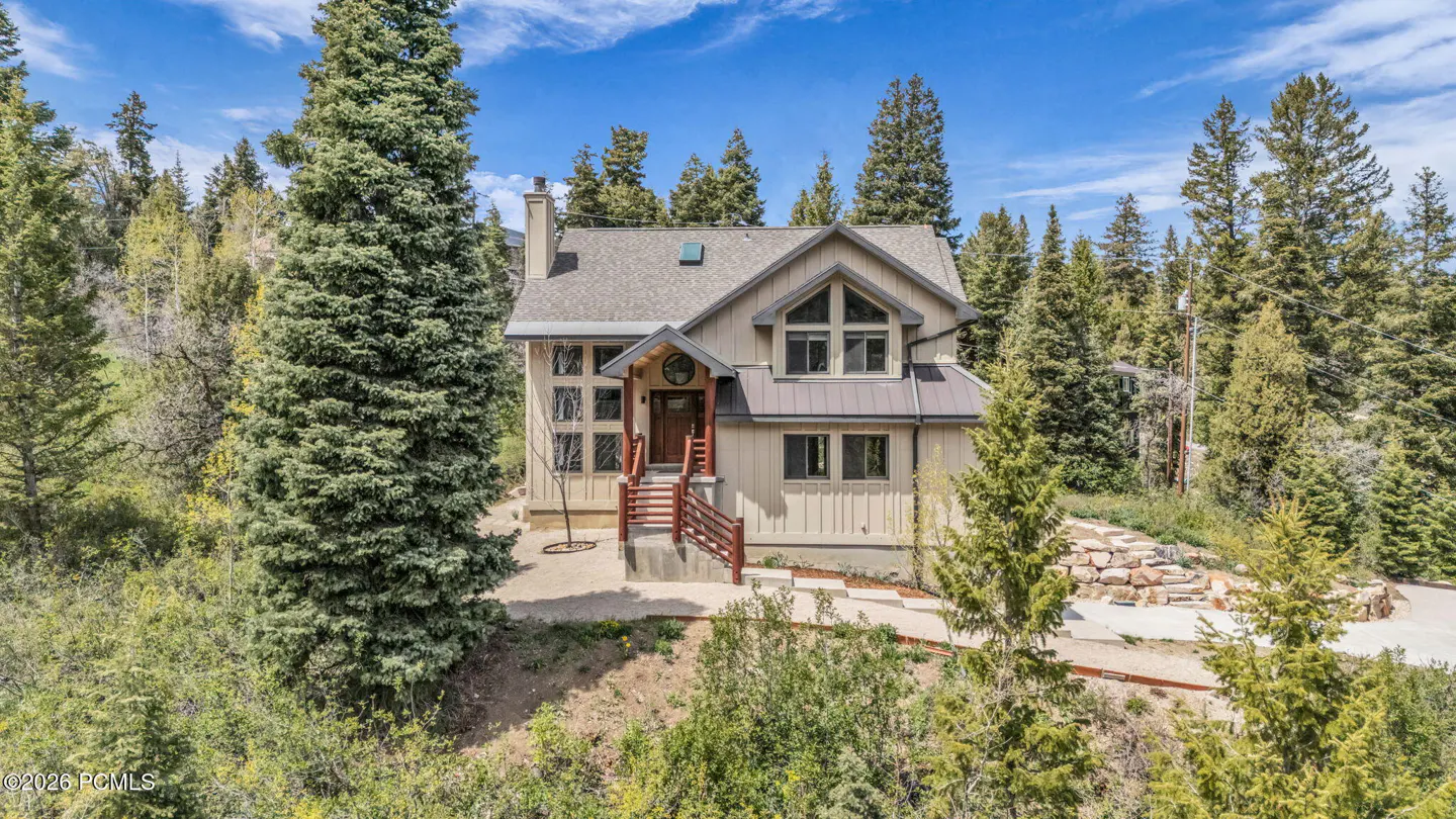 A beige two-story house with a gray roof is surrounded by tall green trees under a blue sky. A wooden staircase leads to the front door.