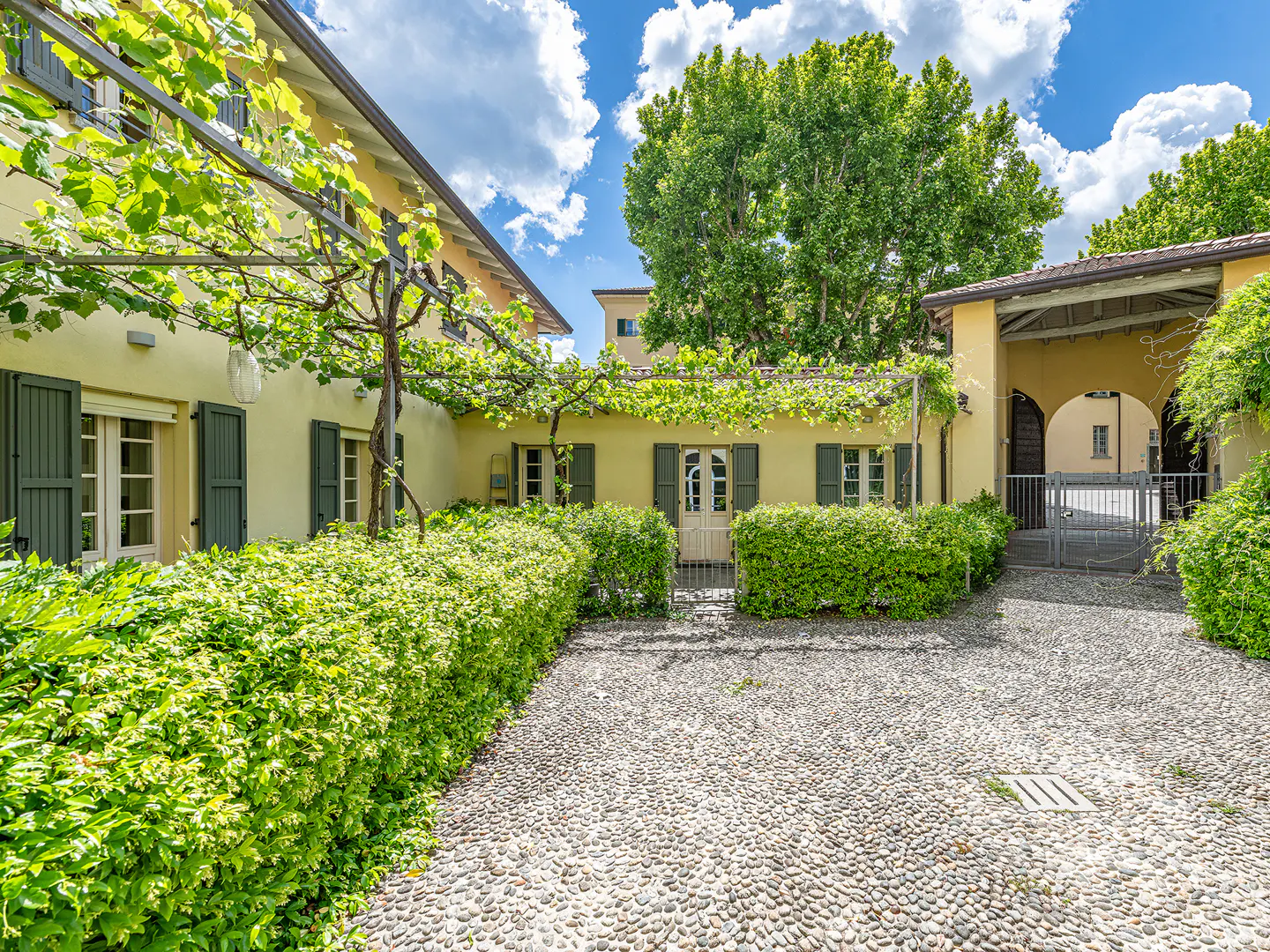 Exterior view of a yellow building with green shutters, a stone courtyard, and lush greenery.