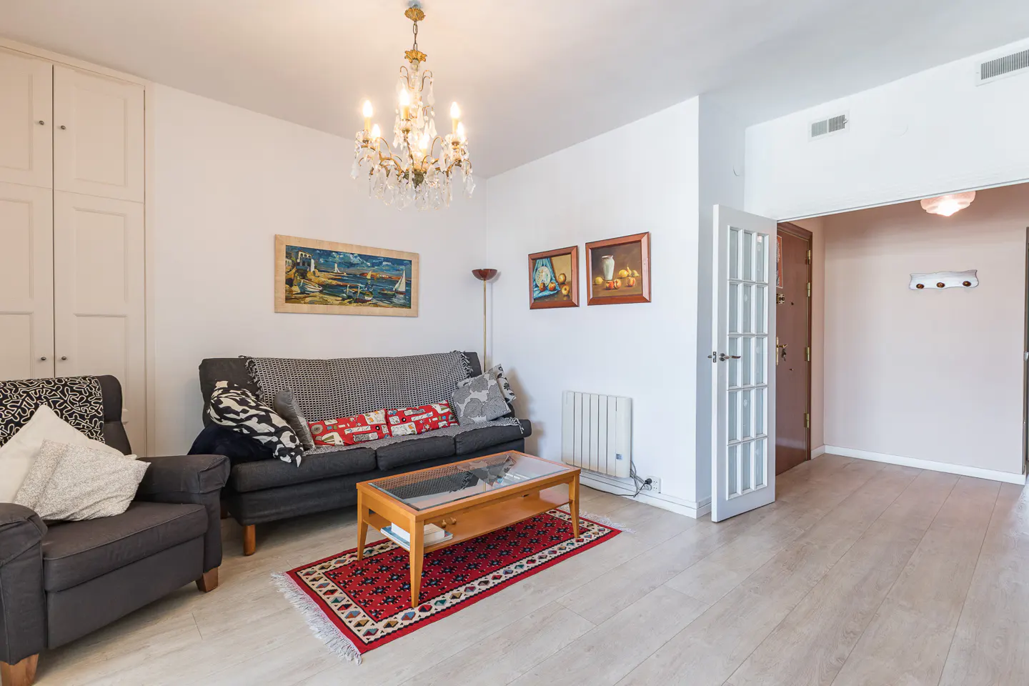 Living room with gray sofa, armchair, and wood coffee table on a red rug. White walls, paintings, and a chandelier.