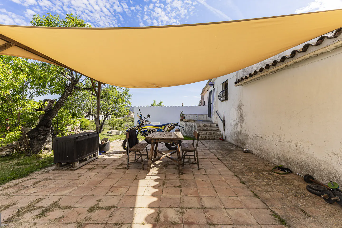 Outdoor patio with a tan sunshade, table, chairs, and a motorcycle parked in the background.