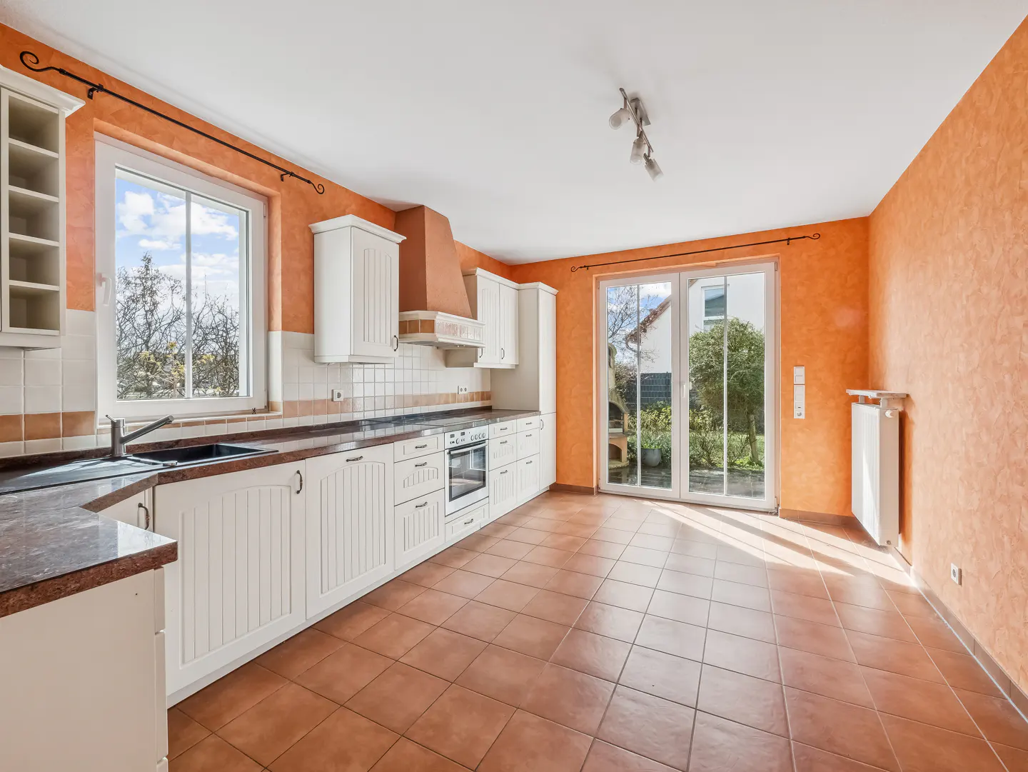 Bright kitchen with orange walls, white cabinets, and brown tile floor. Sliding glass doors lead to a garden.