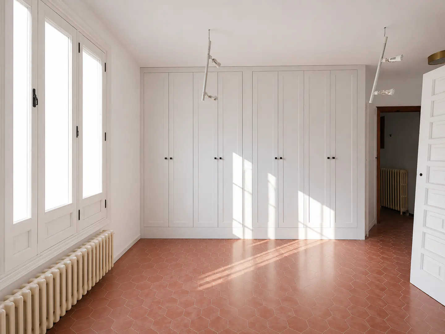 Bright, empty room with white walls, terracotta tile floor, and built-in white wardrobes. Sunlight streams in from a large window.