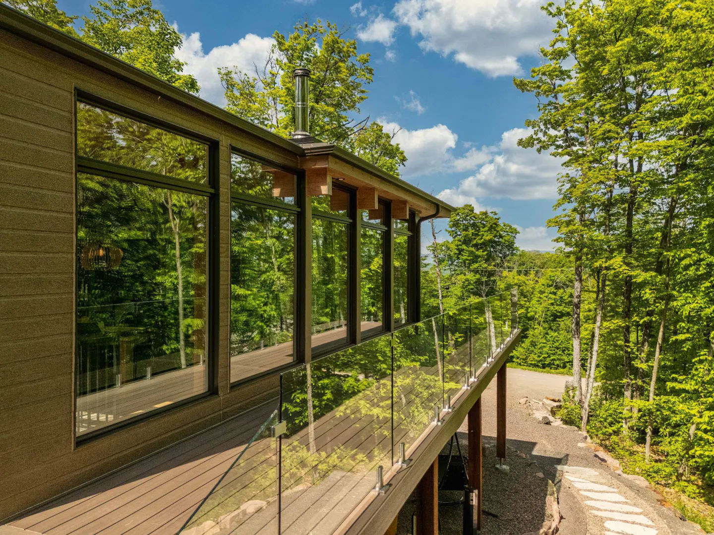Modern home exterior with a wooden deck, glass railing, and large windows reflecting the surrounding lush green forest.