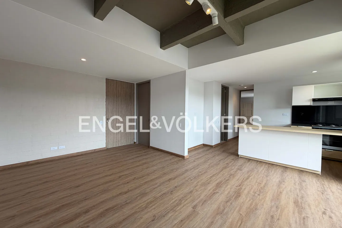 Open-concept living space with wood floors, white walls, and a kitchen island. Gray ceiling beams and track lighting add modern flair.
