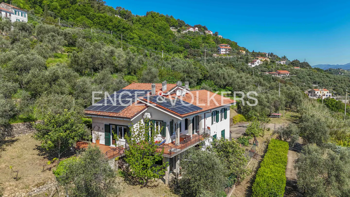 Aerial view of a white two-story house with a red tile roof and solar panels, nestled on a green hillside under a blue sky.