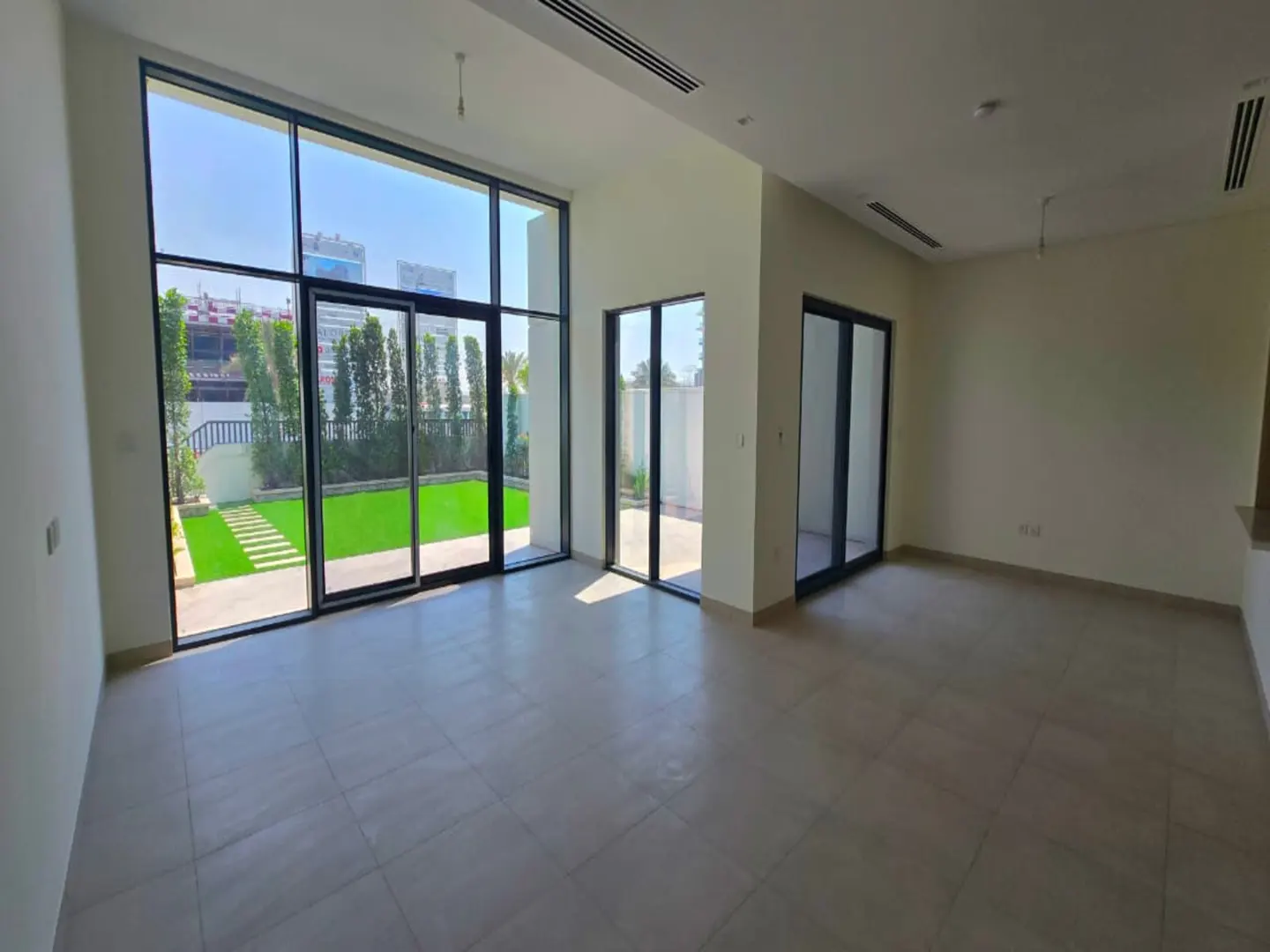 Bright, empty living room with large floor-to-ceiling windows overlooking a green backyard and modern city buildings.