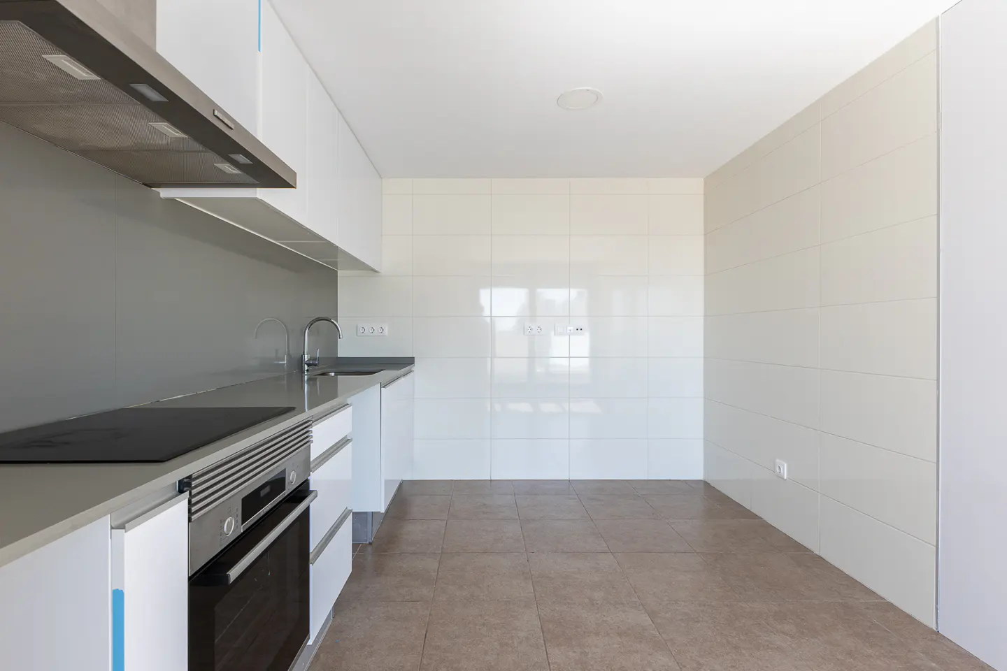 Bright, modern kitchen with white cabinets, gray countertops, stainless steel oven, and brown tile floor.