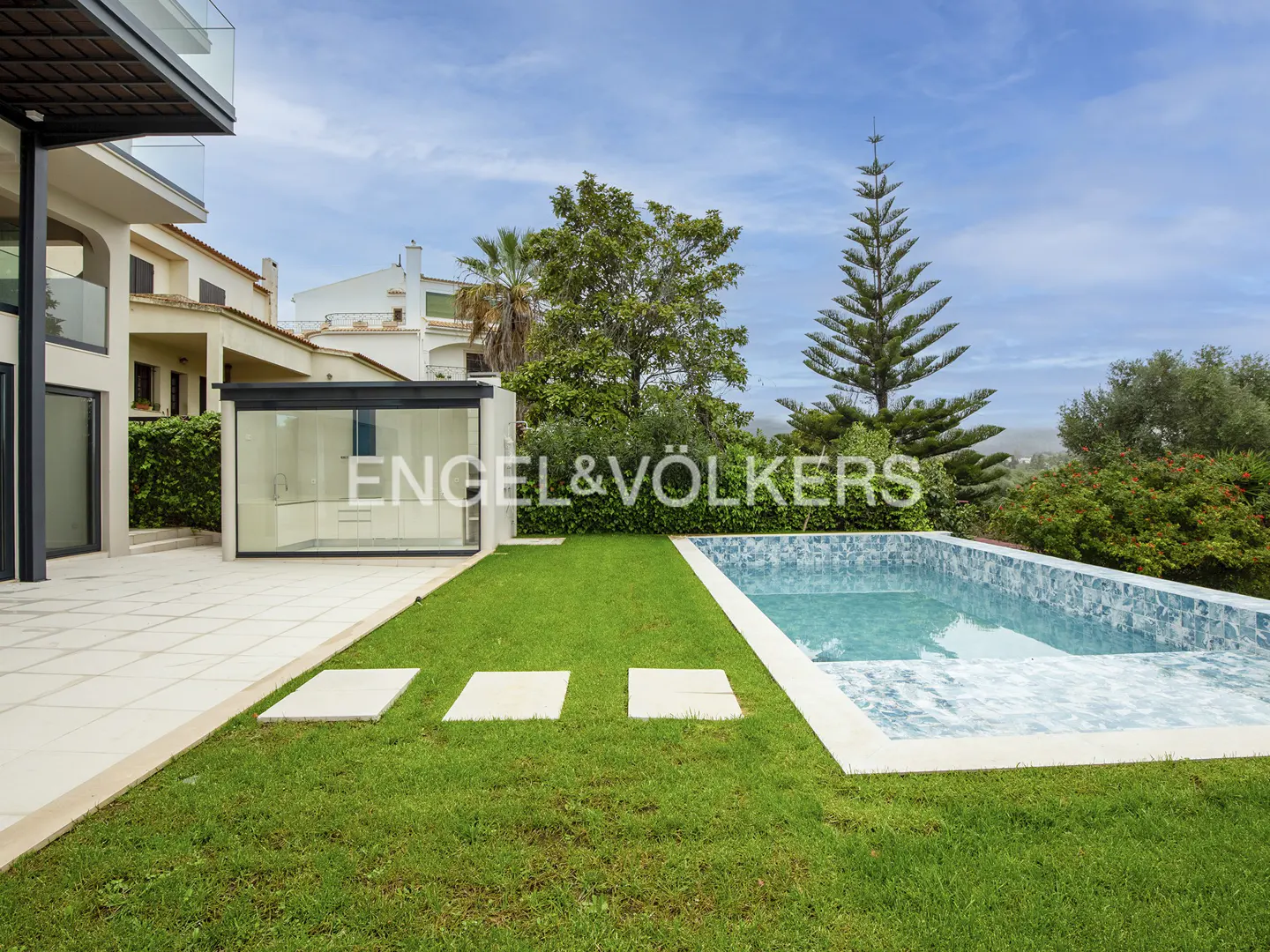 A backyard with a pool, green grass, and a modern house with a glass-enclosed kitchen extension. Trees and blue sky in the background.