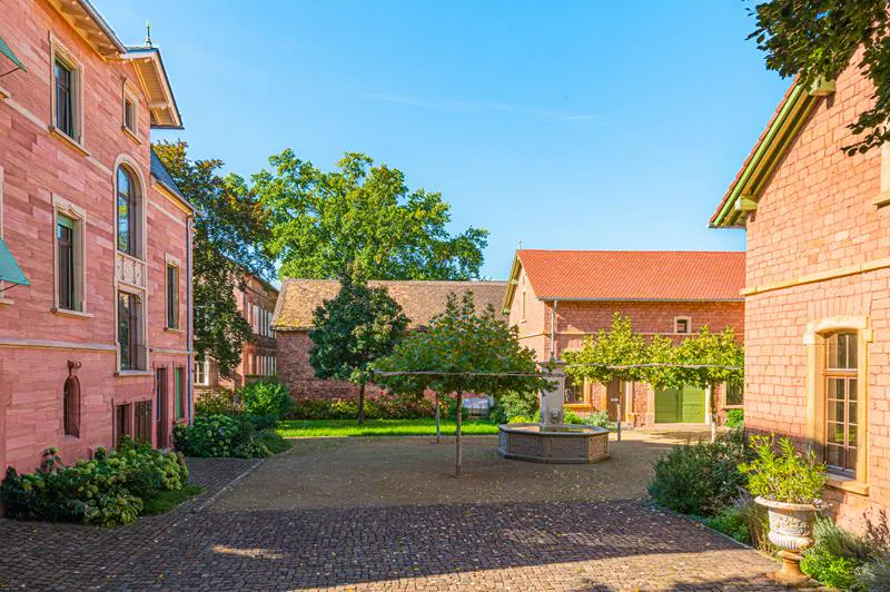Courtyard view of red brick buildings, trees, and a stone fountain under a clear blue sky.