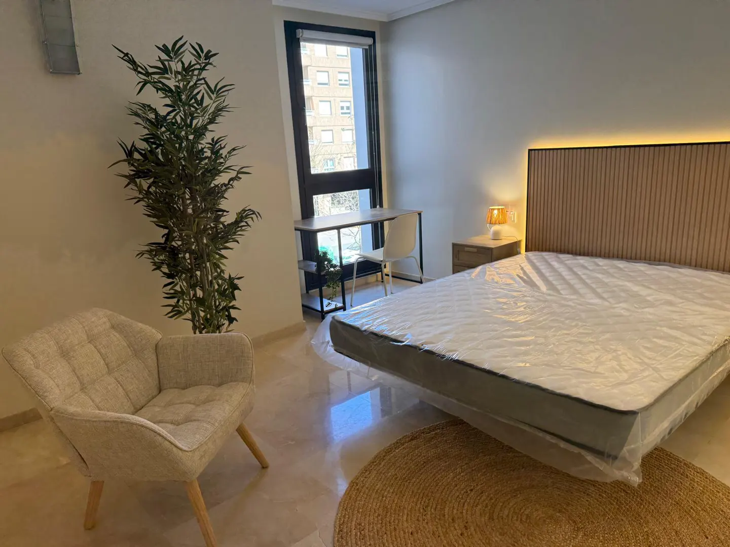 Bedroom with a bed covered in plastic, a desk by the window, a chair, and a plant.