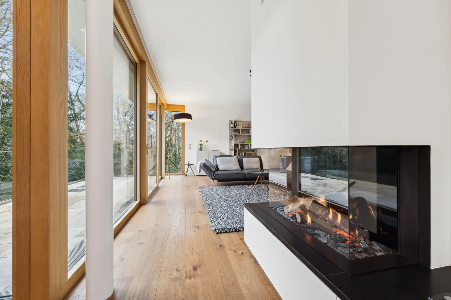 Bright living room with wood floors, a modern fireplace, and floor-to-ceiling windows overlooking trees. A black sofa sits on a gray rug.