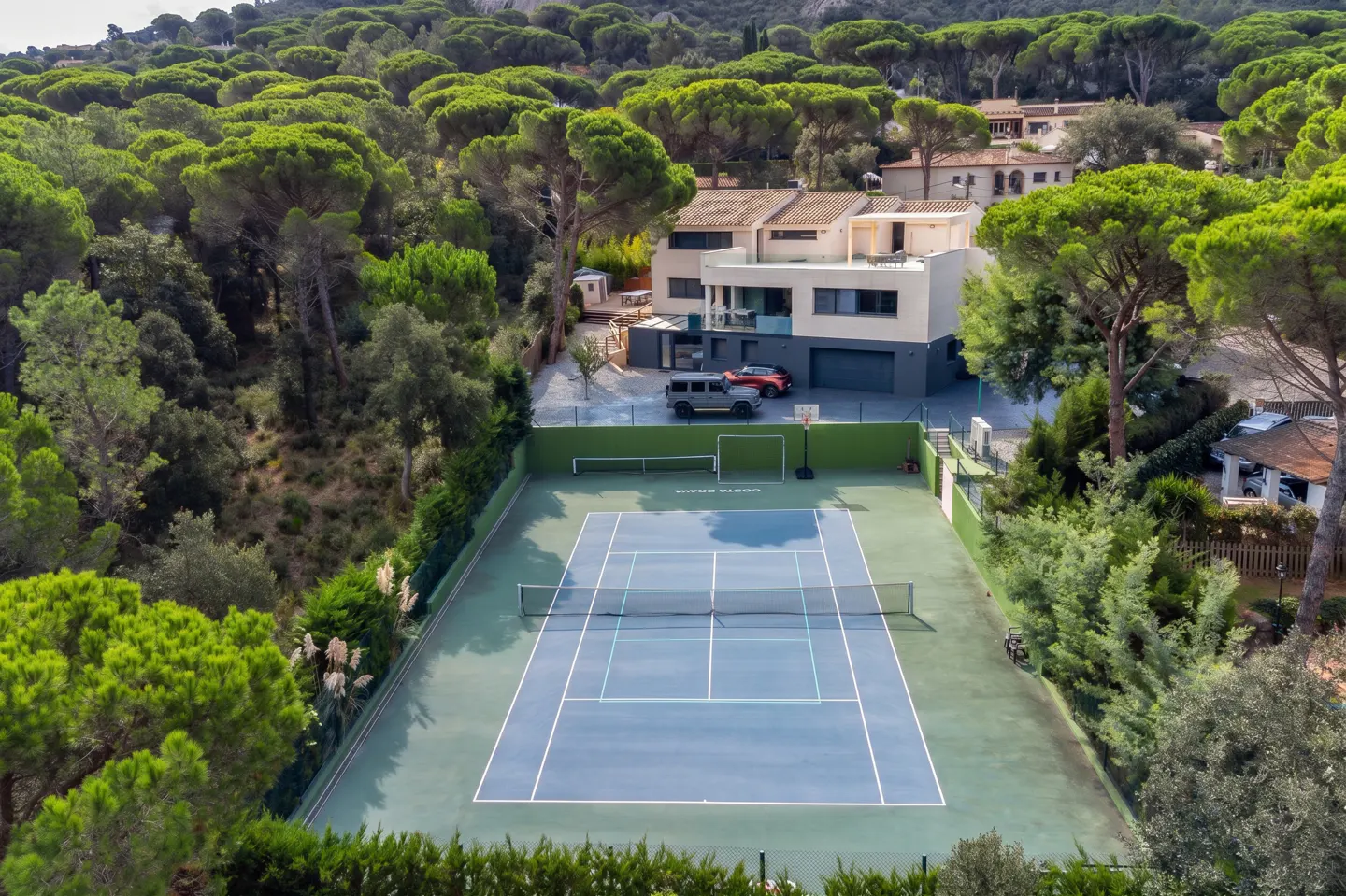 Aerial view of a modern home with a tennis court, surrounded by lush green trees. Two cars are parked near the house.