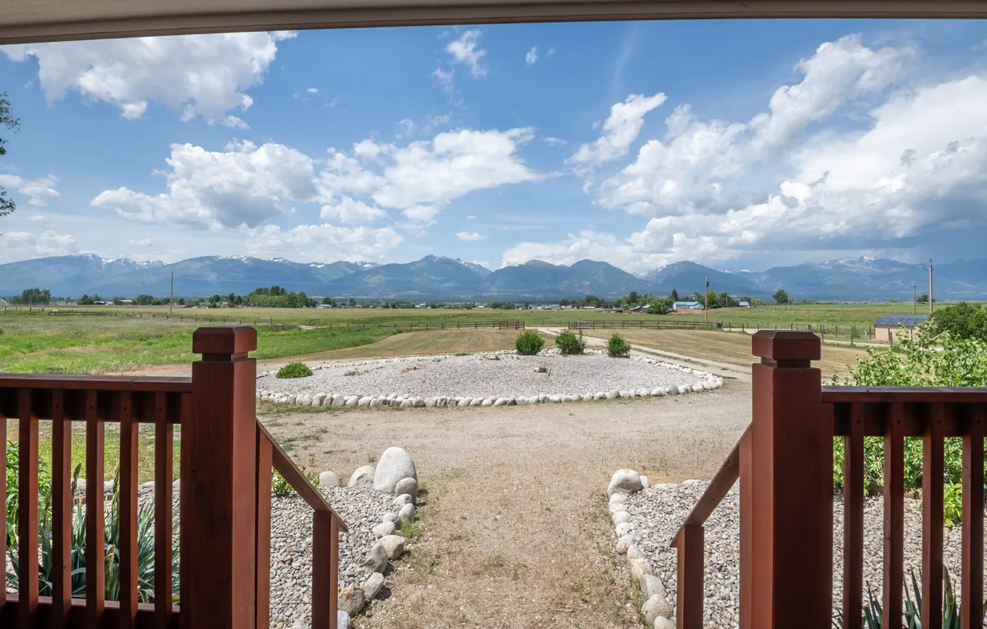 View from a porch with brown railings, overlooking a gravel yard, green fields, and distant mountains under a blue, cloudy sky.