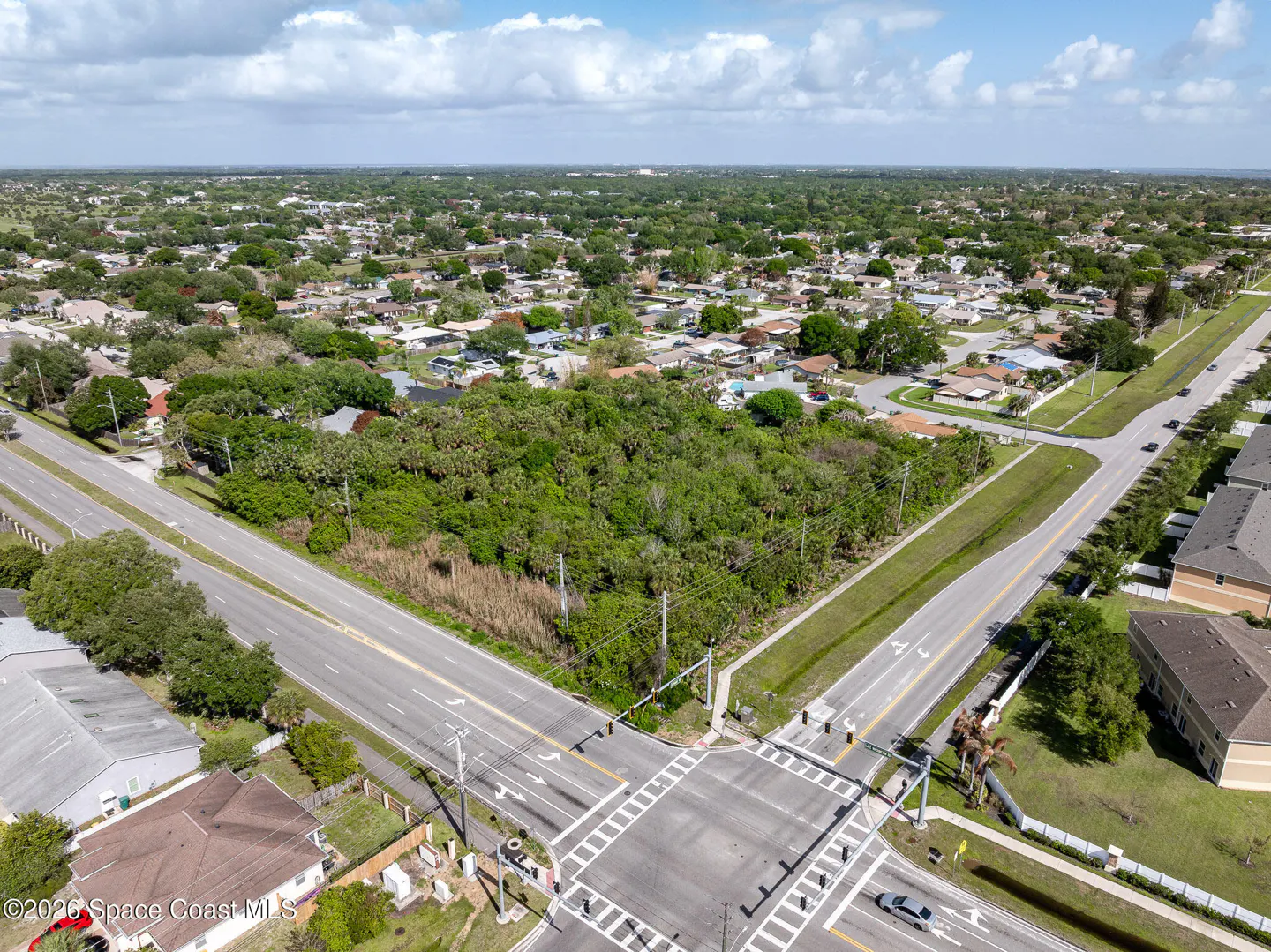 Aerial view of a wooded lot at a road intersection in a residential area under a cloudy sky.