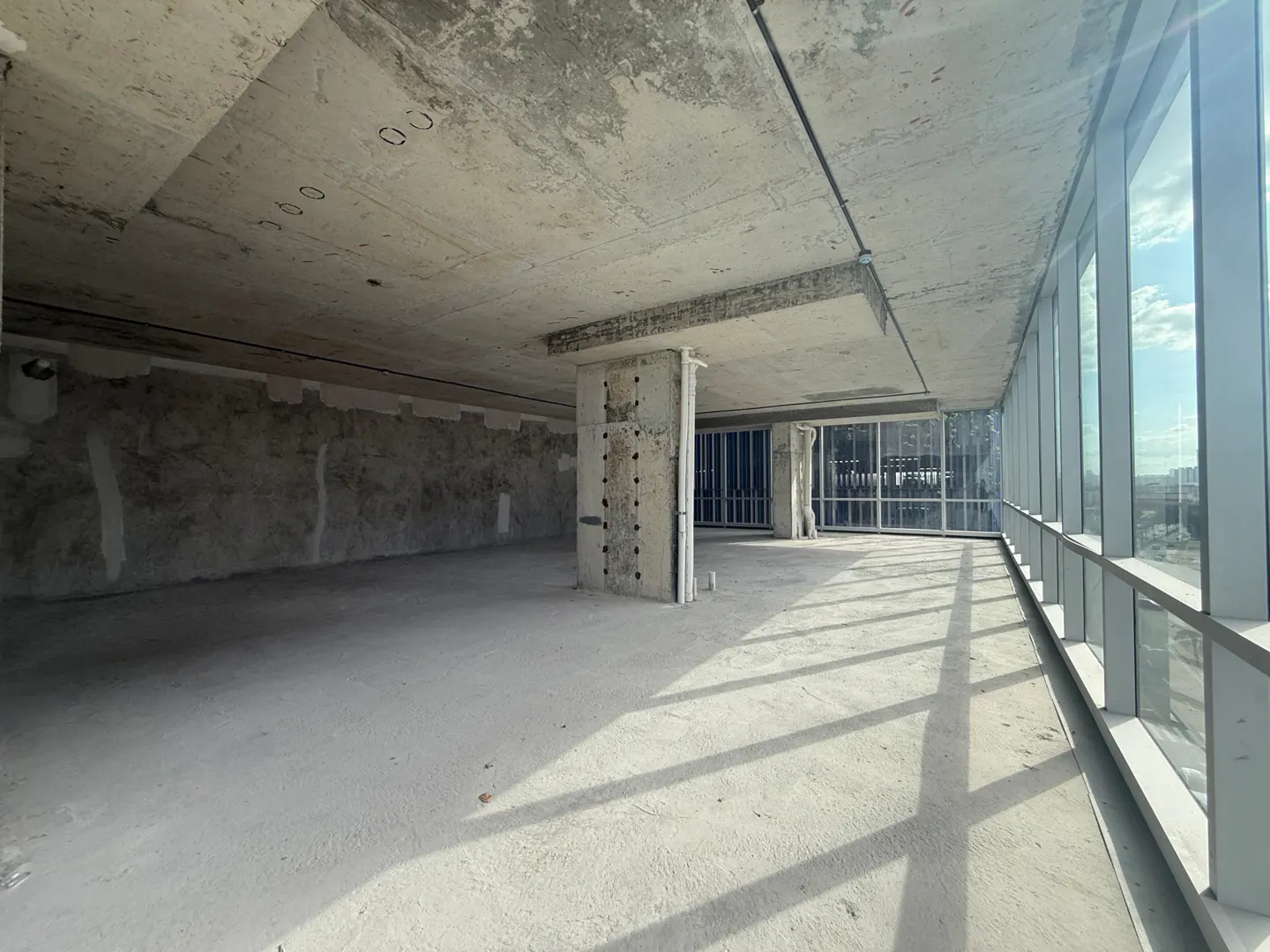 Unfinished gray concrete room with floor-to-ceiling windows. Sunlight casts shadows across the floor.
