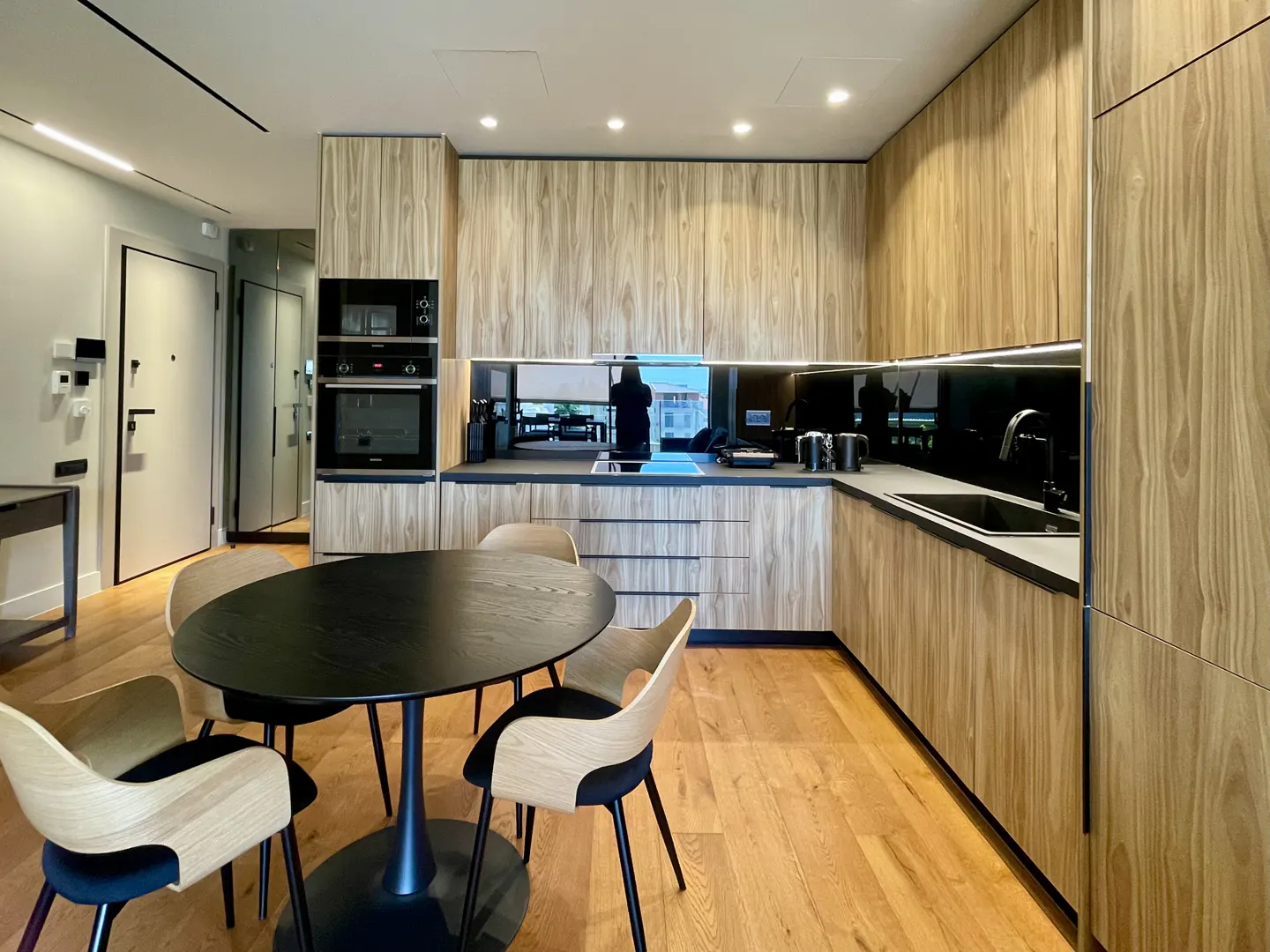 A modern kitchen with wood-grain cabinets, black countertops, and a round black table with four chairs.
