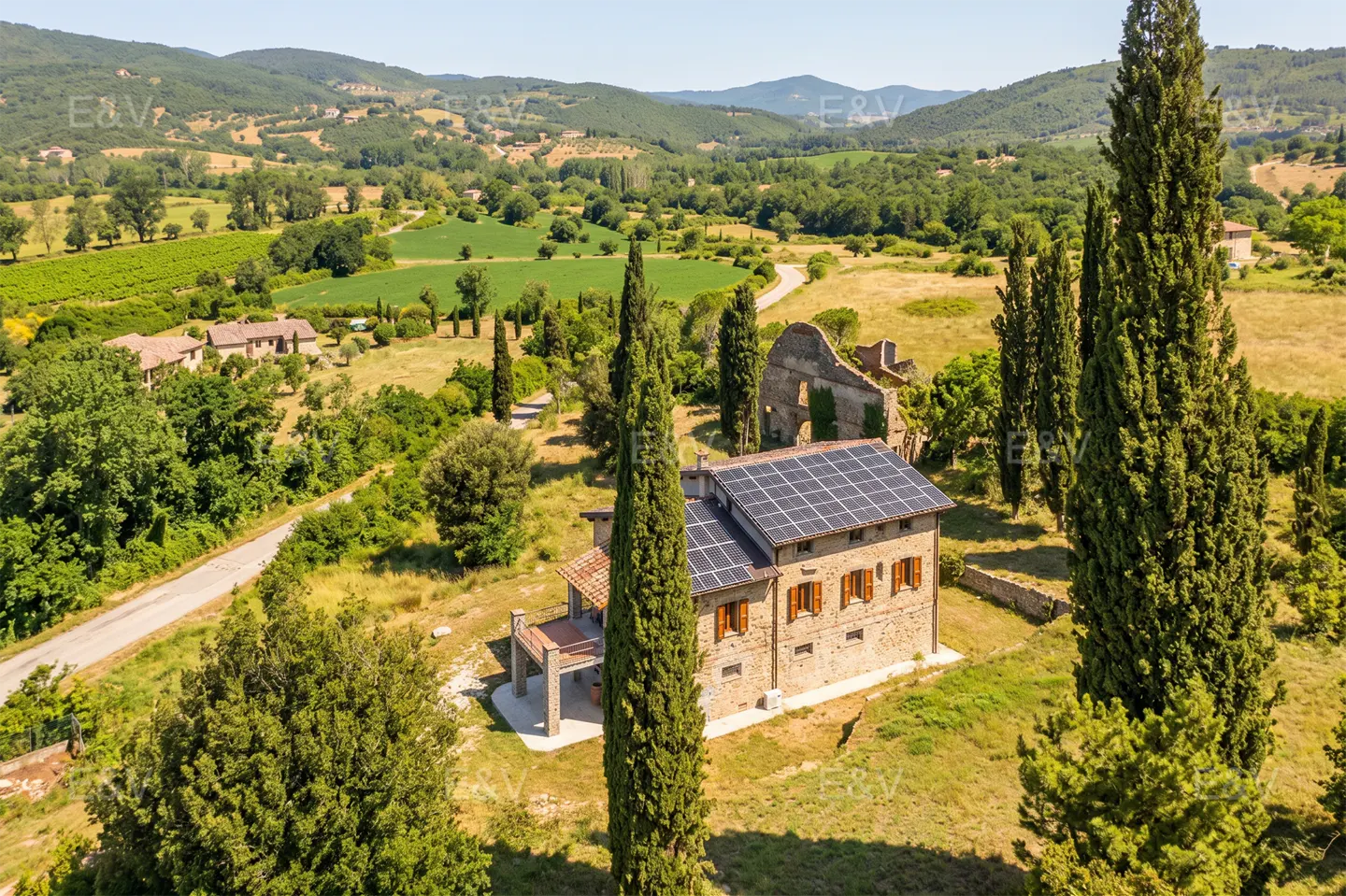Aerial view of a stone house with solar panels, brown shutters, and tall cypress trees in a green, hilly landscape.