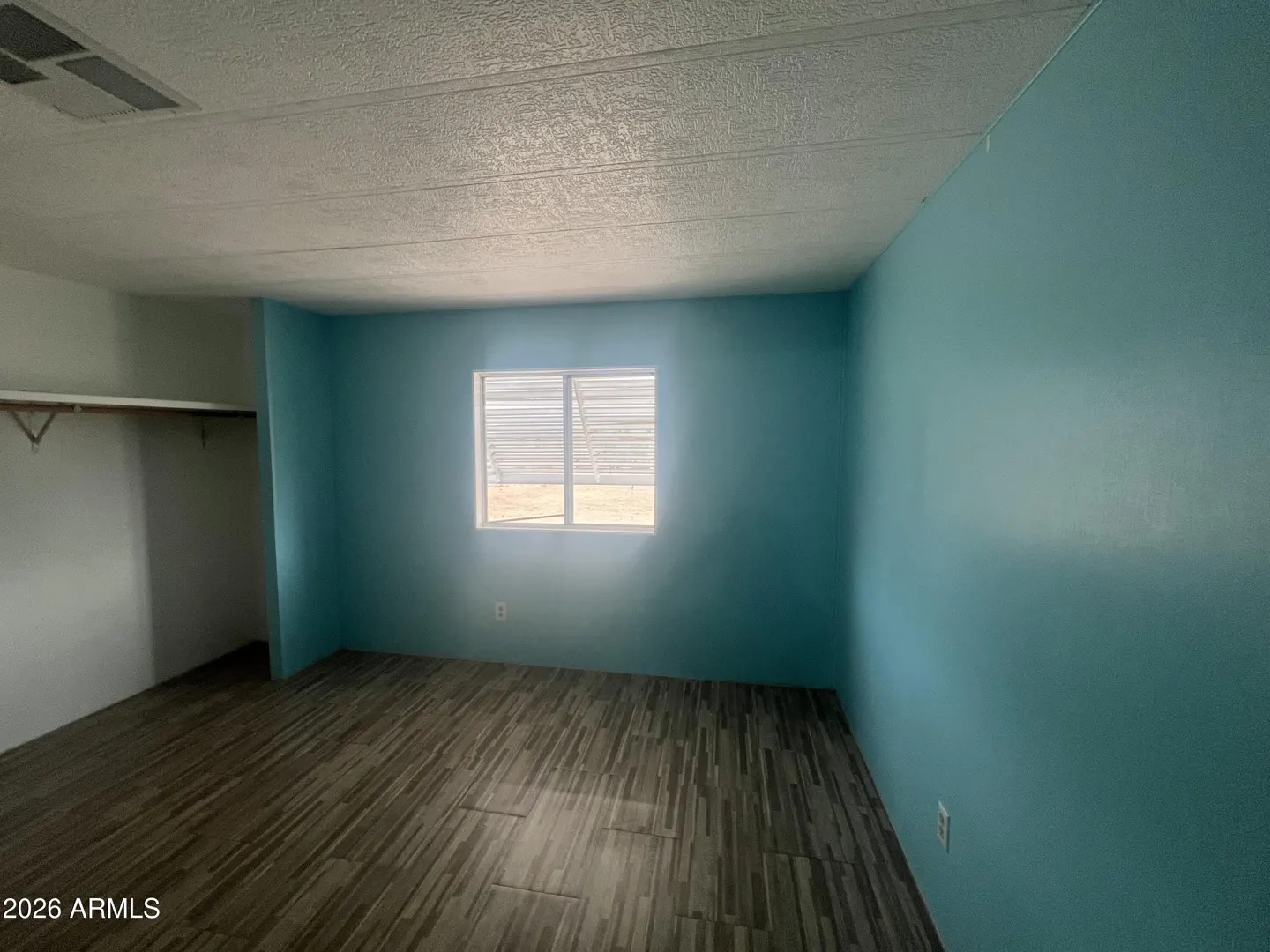 Empty bedroom with light blue walls, wood-look tile floor, and a window letting in natural light. A closet is visible on the left.