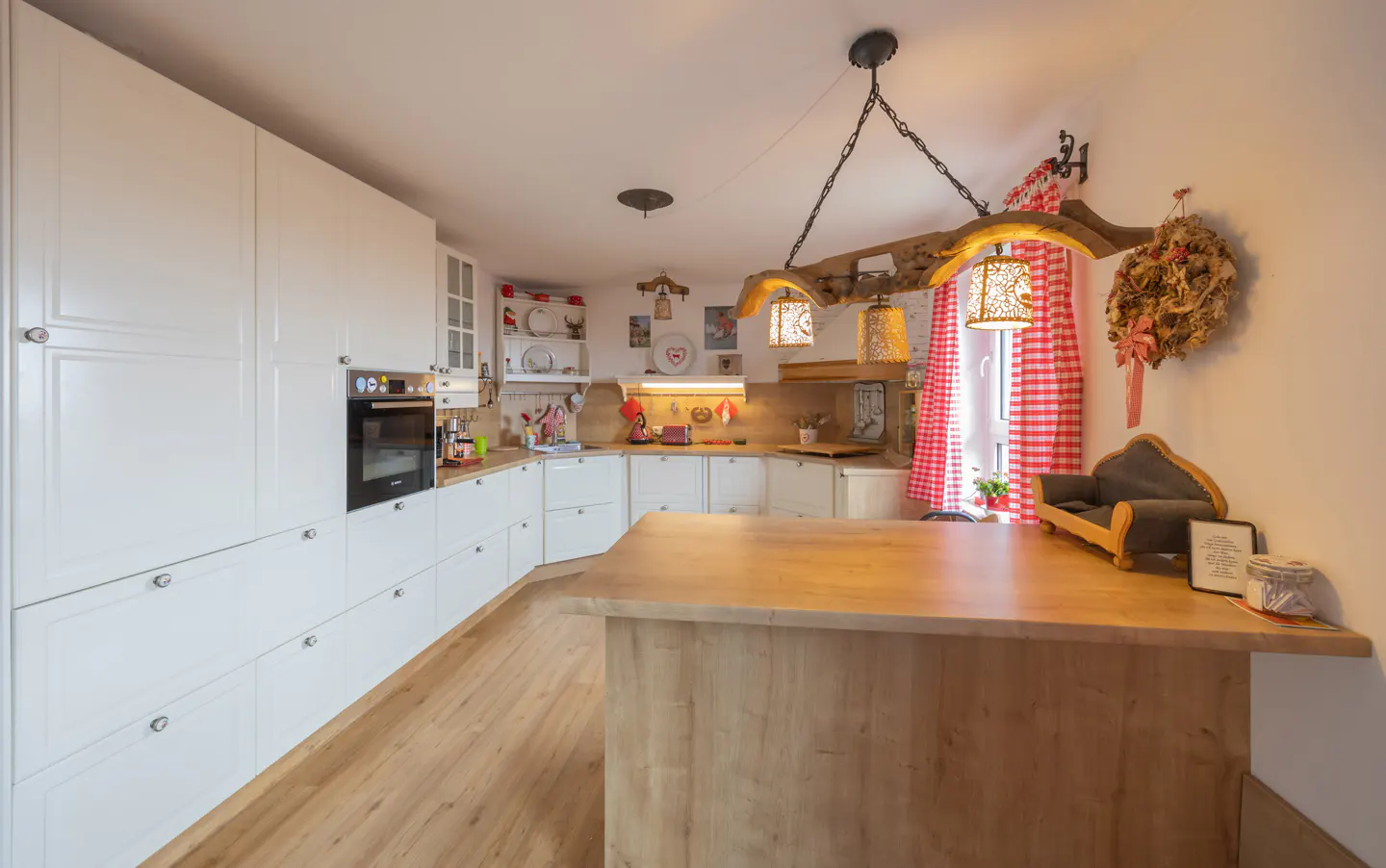 A bright kitchen with white cabinets, wood floors, and a large wooden island. A rustic chandelier hangs above the island. Red and white checkered curtains are on the window.