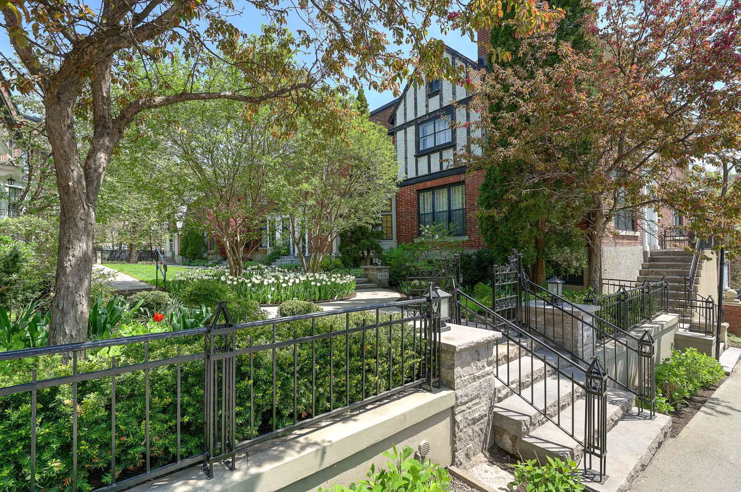 Exterior view of a brick and stucco Tudor-style house with a black iron fence and stone steps leading to the entrance. Lush greenery and trees surround the property.