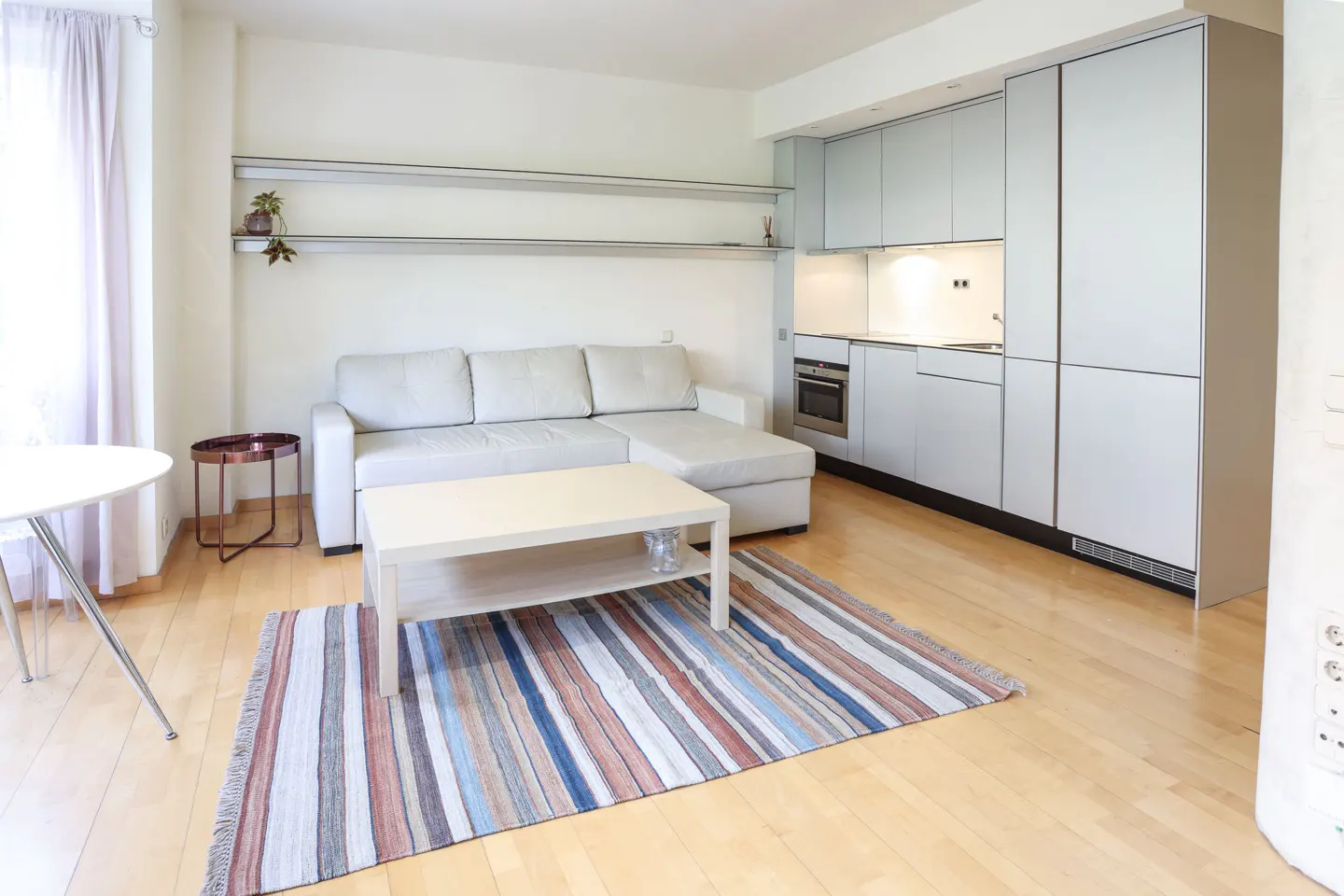 A bright studio apartment with a white couch, light wood floors, and a modern kitchen. A striped rug adds color to the space.