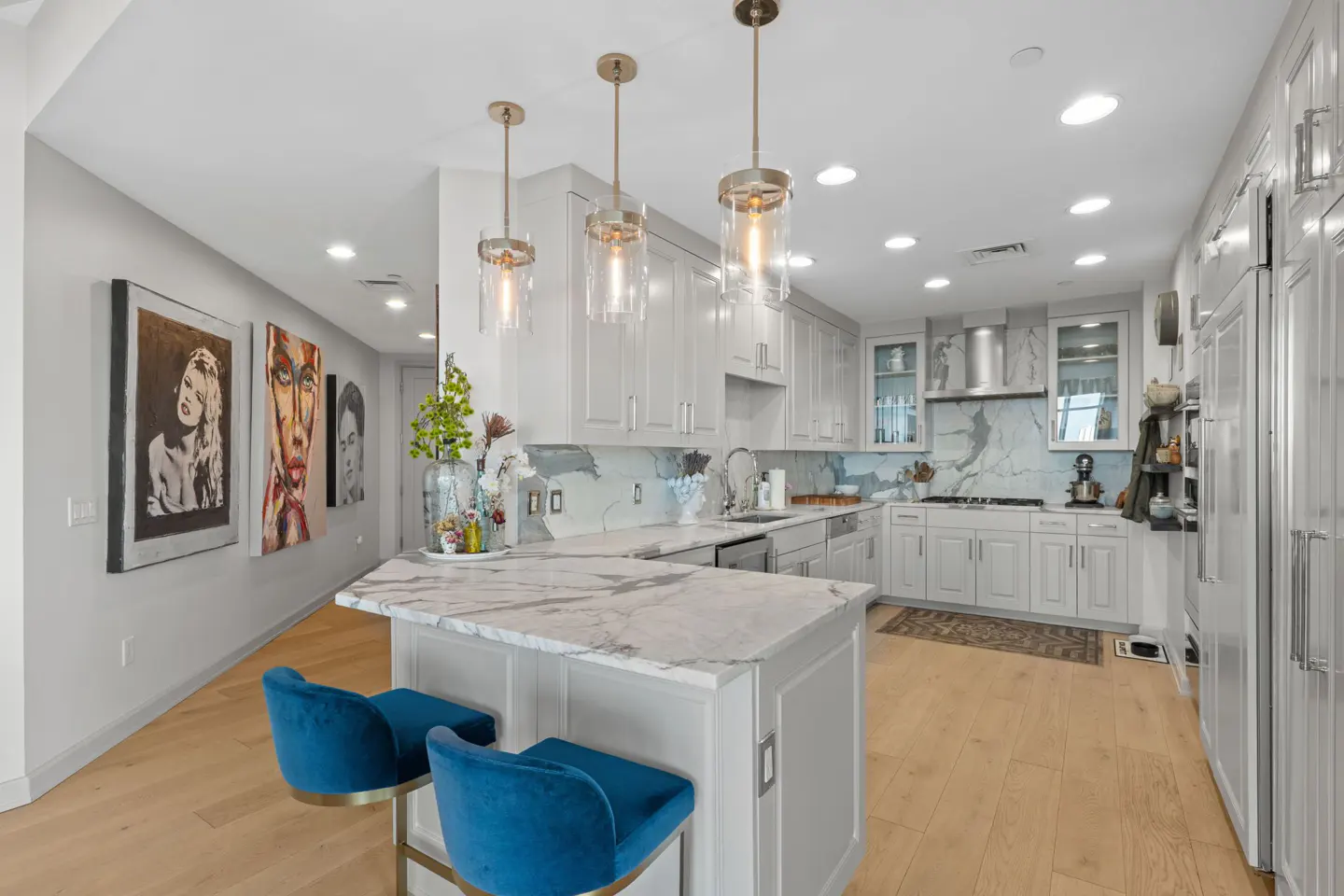 Bright kitchen with white cabinets, marble countertops, and wood floors. Blue velvet bar stools sit at the island. Artwork hangs on the wall.