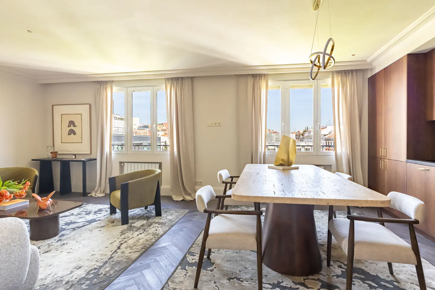 Modern dining and living area with a stone table, cream chairs, olive armchairs, and large windows overlooking a city.
