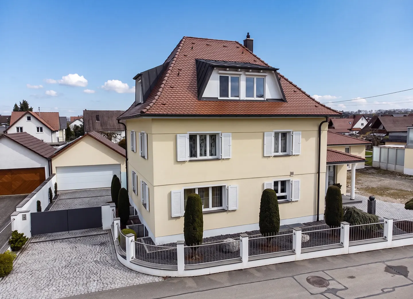 Two-story yellow house with a red tile roof and white shutters, surrounded by a white fence and green shrubbery.