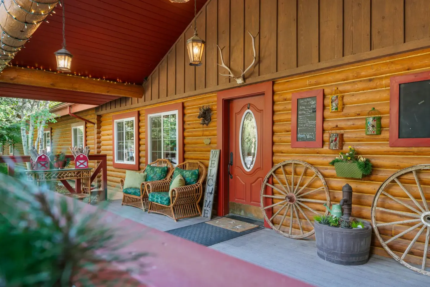 Rustic log cabin porch with red door, wicker chairs, wagon wheels, and decorative wall hangings under a covered roof.