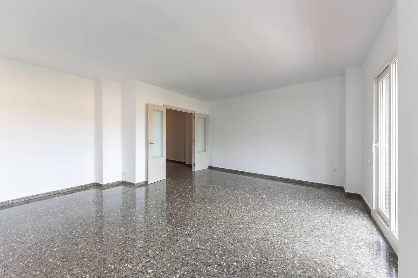 Empty room with white walls, grey terrazzo flooring, and open double doors leading to a hallway.